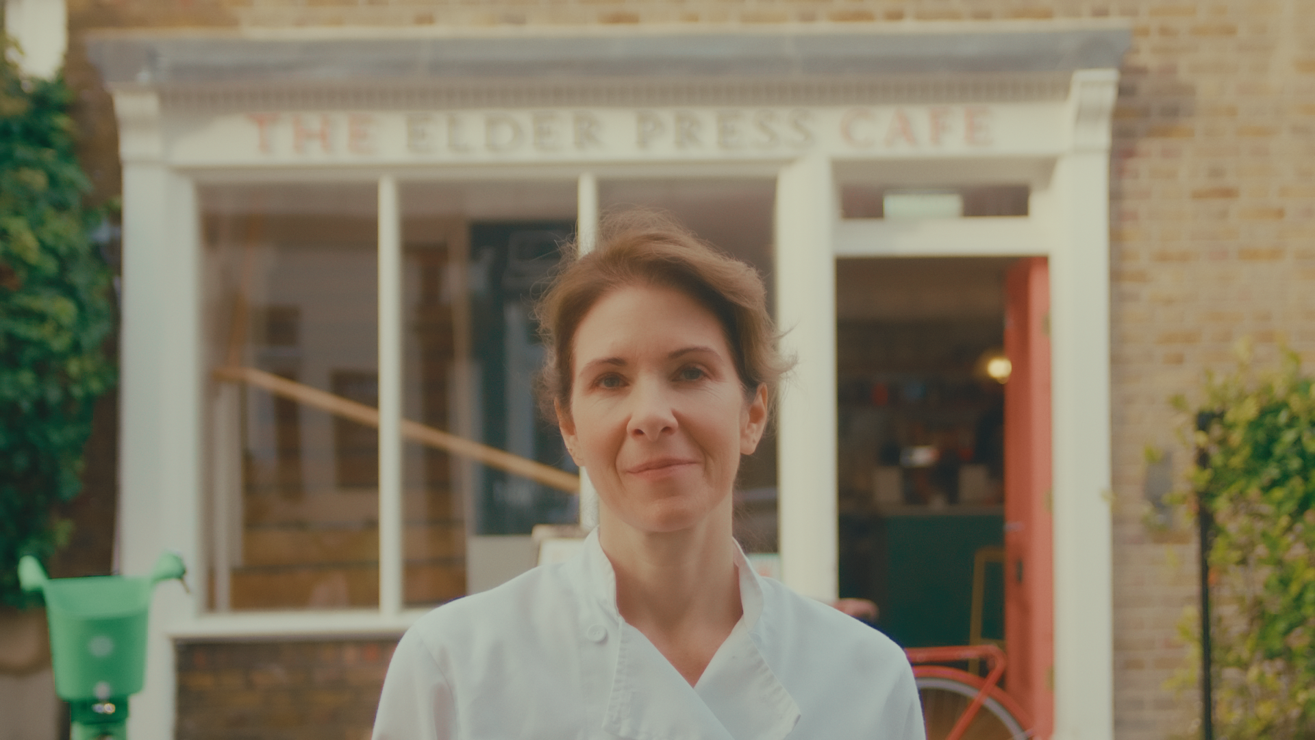 A woman in a chef's coat standing outside of a café called The Elder Press Cafe, with a green bike on her right and greenery on her left.