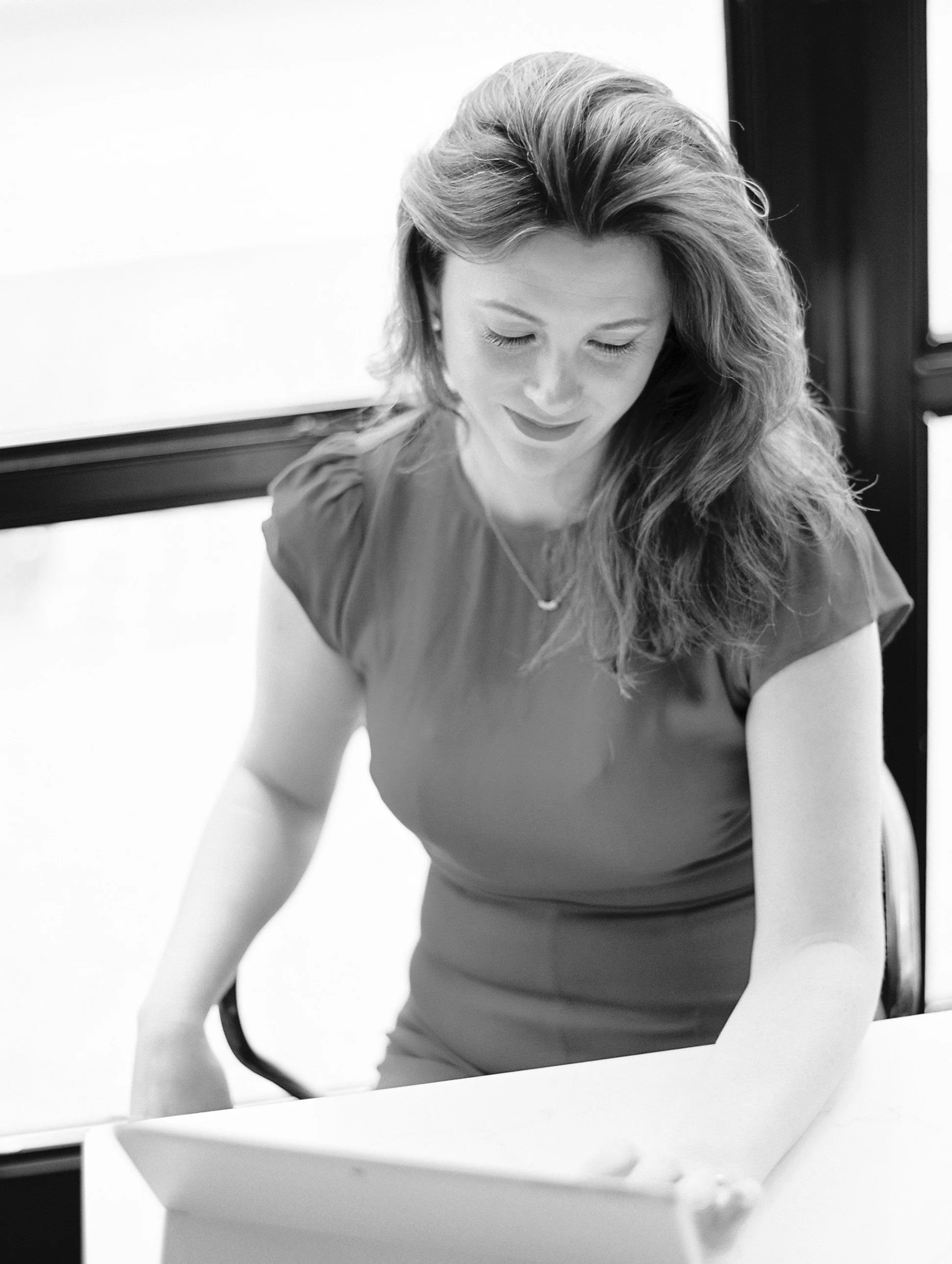 A woman with wavy hair is sitting at a table looking down at a tablet.