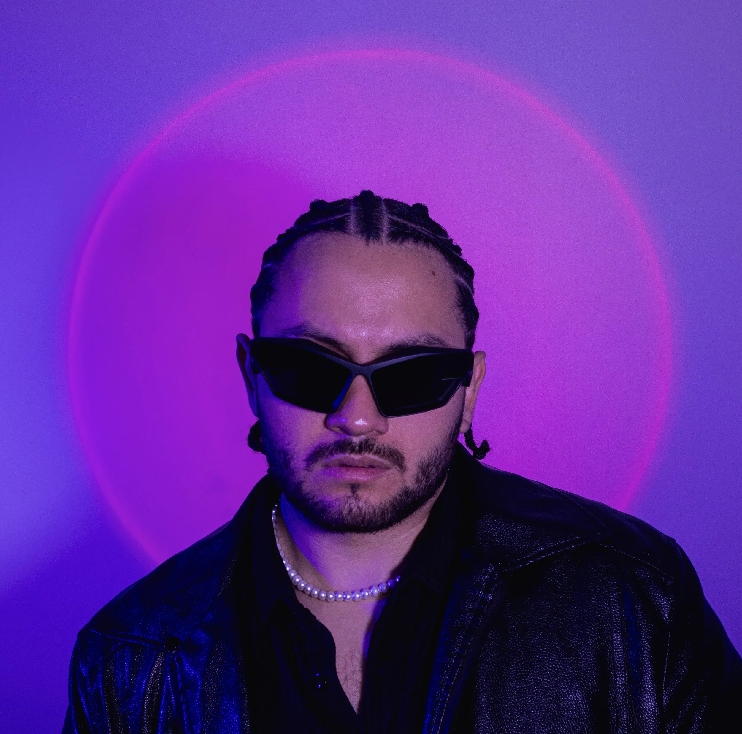 Retrato de un hombre con peinado trenzado y barba, usando gafas de sol negras y un collar de perlas, con fondo morado y rosa brillante.