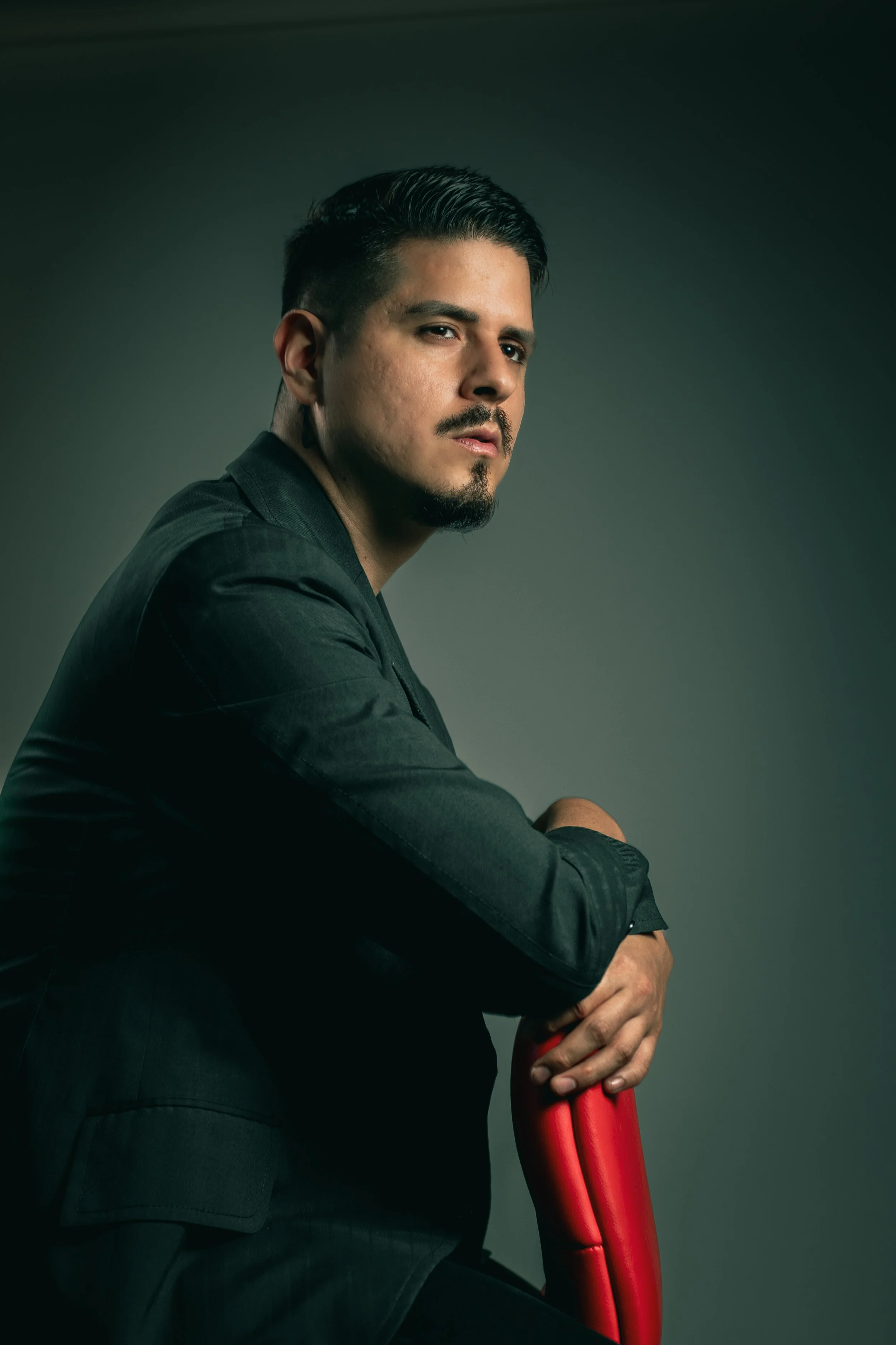 Hombre con barba, bigote y cabello oscuro, usando un saco negro, sentado en una silla roja, con fondo gris oscuro.
