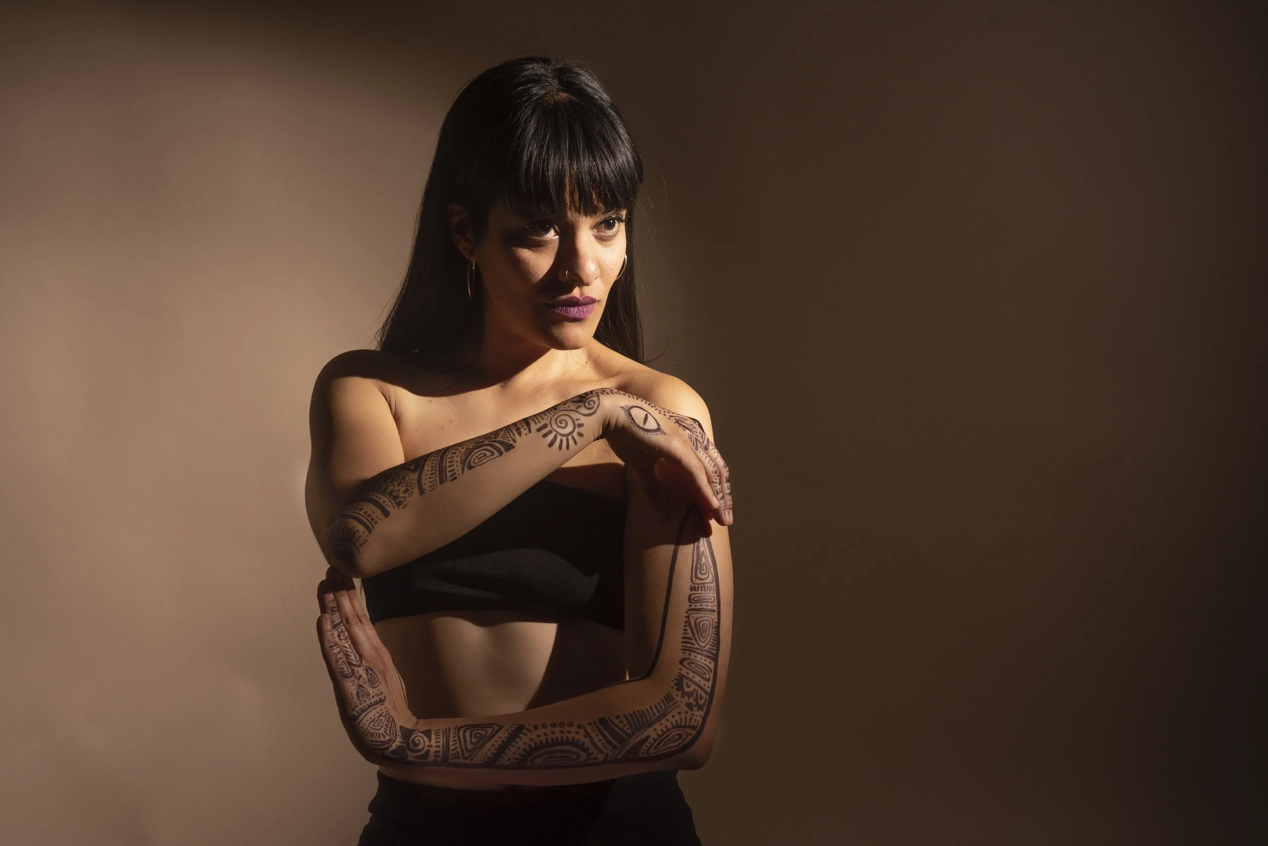Mujer joven con tatuajes tribales en brazos y hombros, con cabello oscuro y flequillo, posa con una expresión pensativa en un fondo de color marrón.