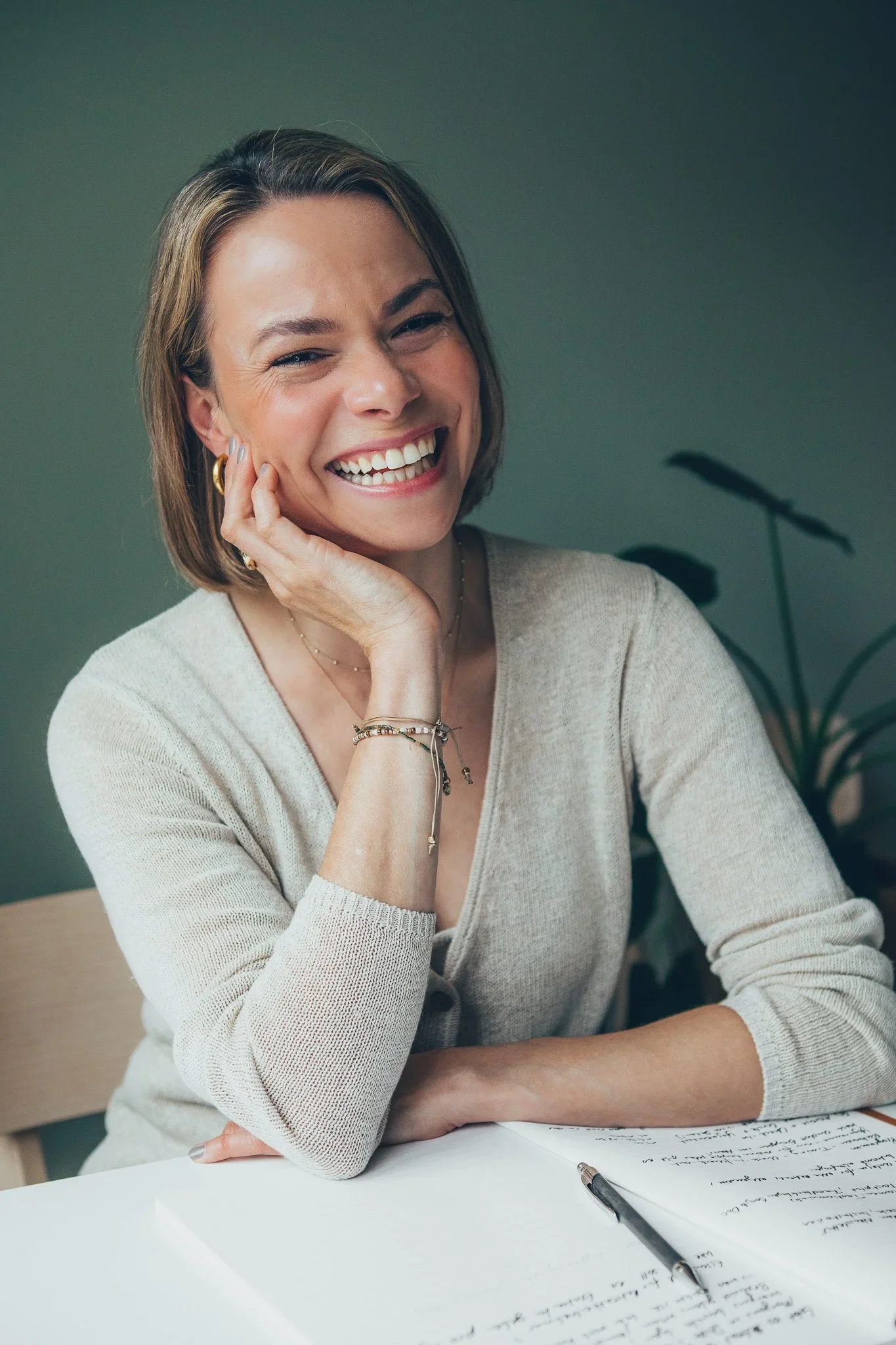 A smiling woman with shoulder-length blonde hair standing against a stone wall. She is wearing a black sleeveless top with thin straps and a watch on her left wrist.