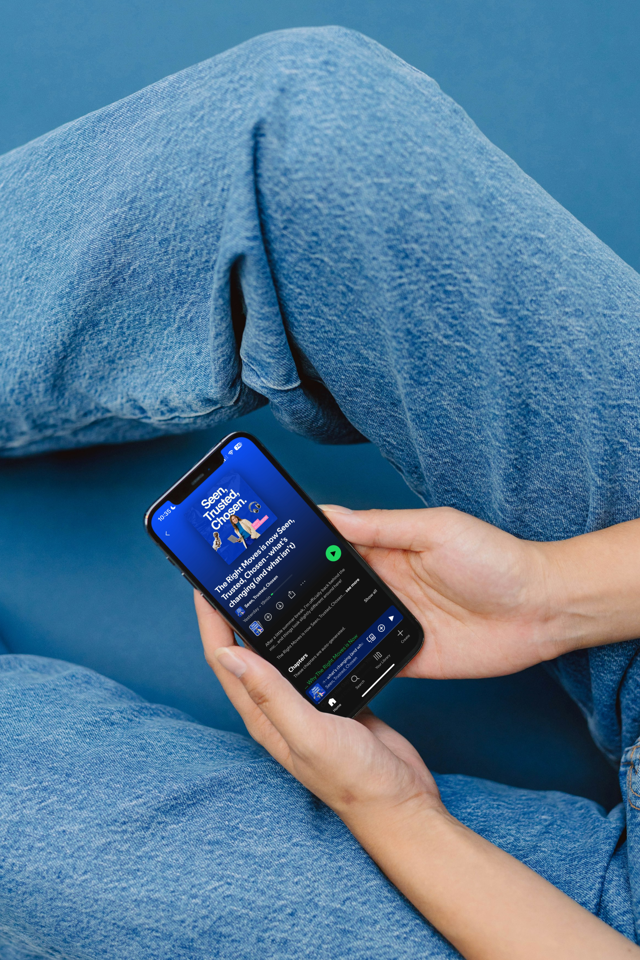 Person sitting cross-legged while holding a smartphone, with focus on the phone screen showing a podcast app playing a podcast episode.