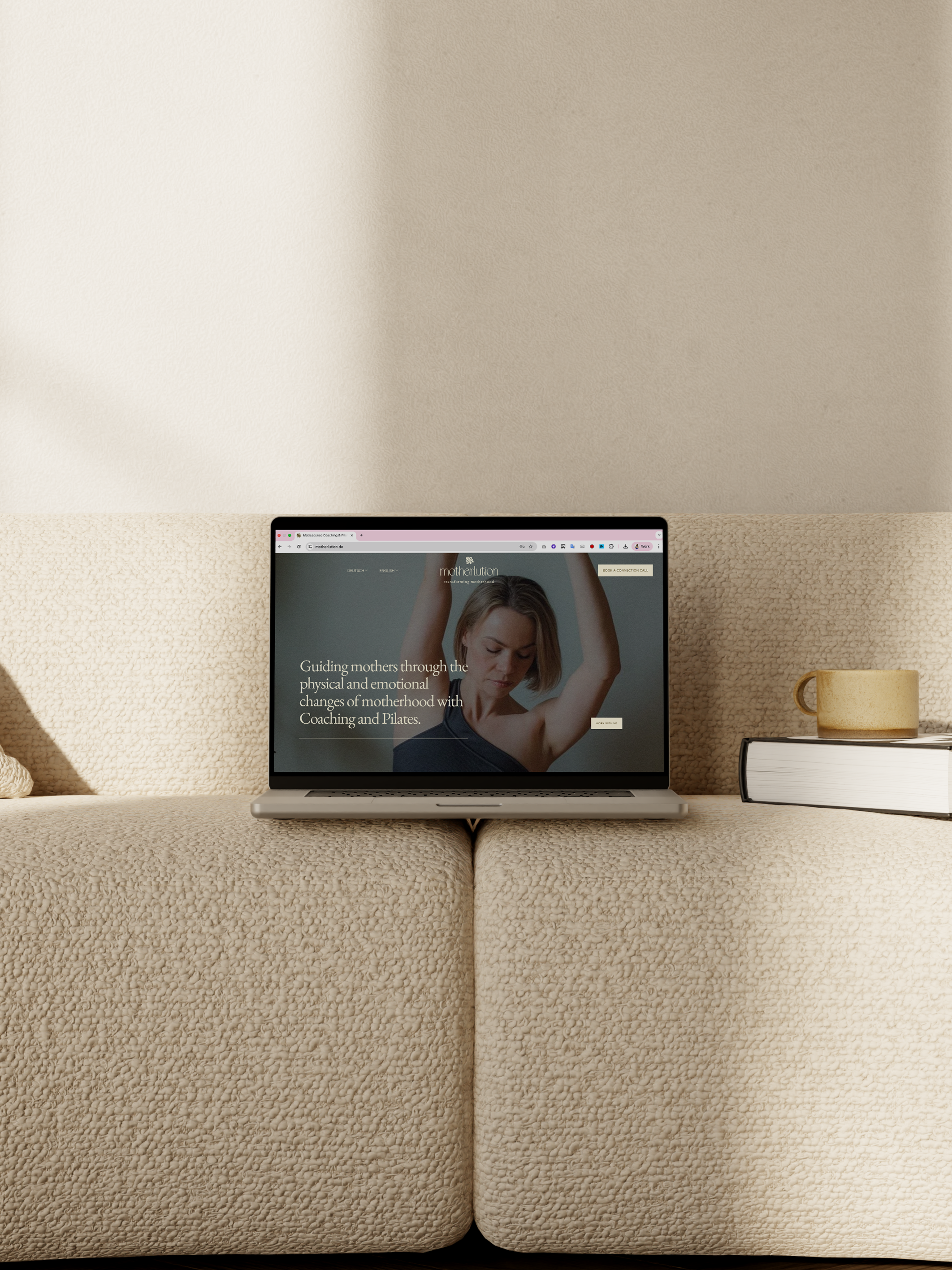 A laptop on a beige, textured sofa, with a yellow mug and a closed book beside it. The laptop screen displays a website about guiding mothers through pregnancy and motherhood.