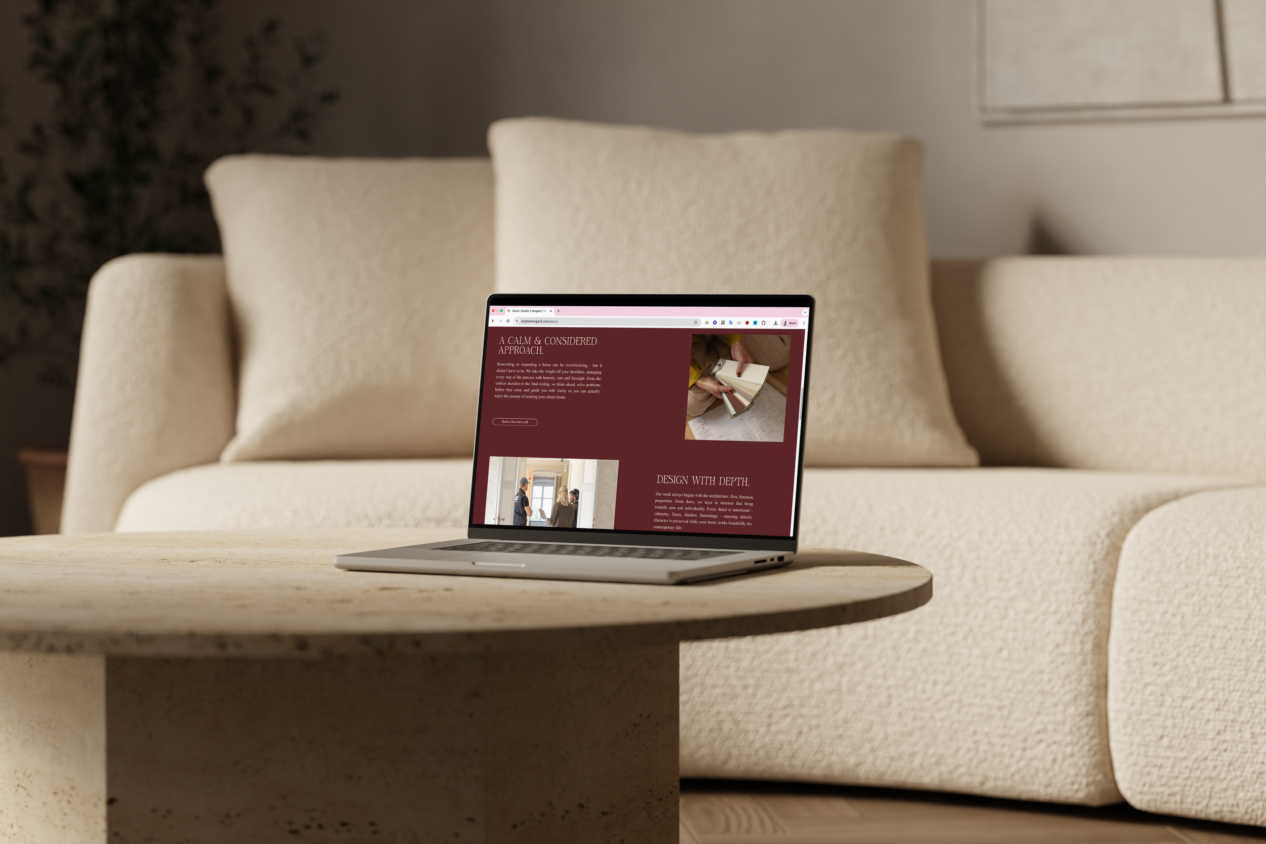 A laptop on a beige, speckled stone table displaying a website with a maroon background and text about home renovation, in front of a cream-colored sofa with large cushions.