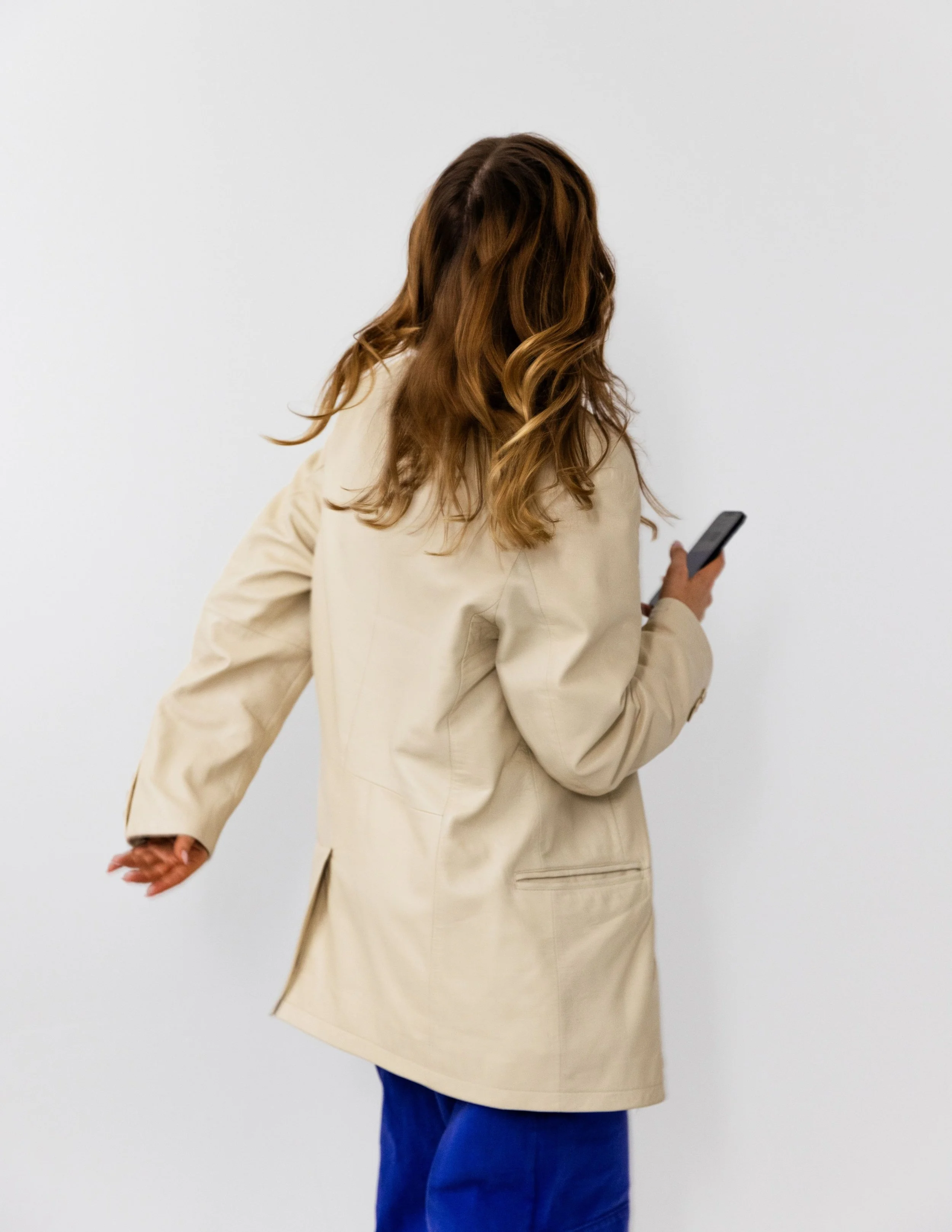 A person with long, wavy hair wearing a beige jacket and blue pants, holding a smartphone, standing against a plain white background.