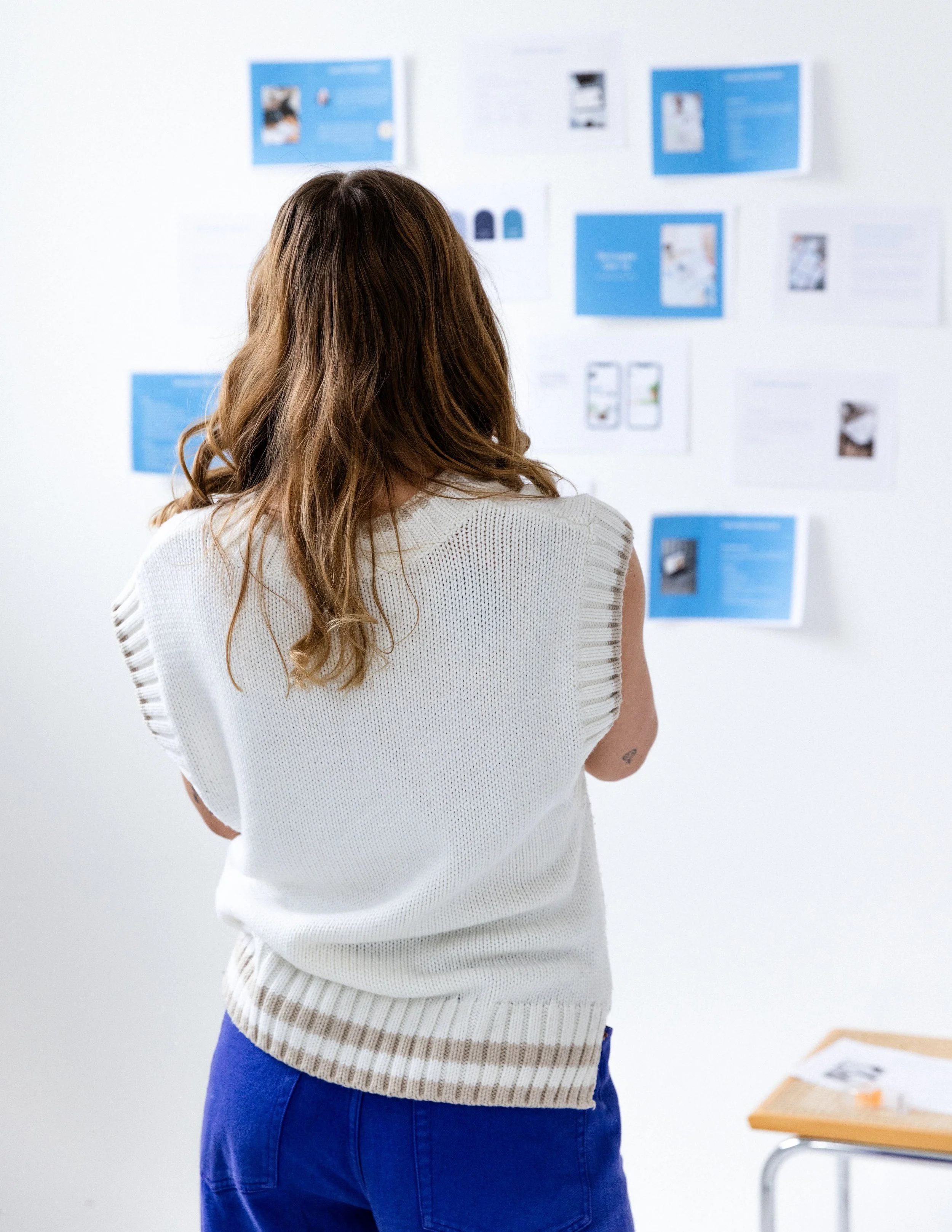 A woman with long brown hair wearing a white knitted sleeveless top and blue skirt is standing in an art gallery, looking at artwork displayed on a white wall.