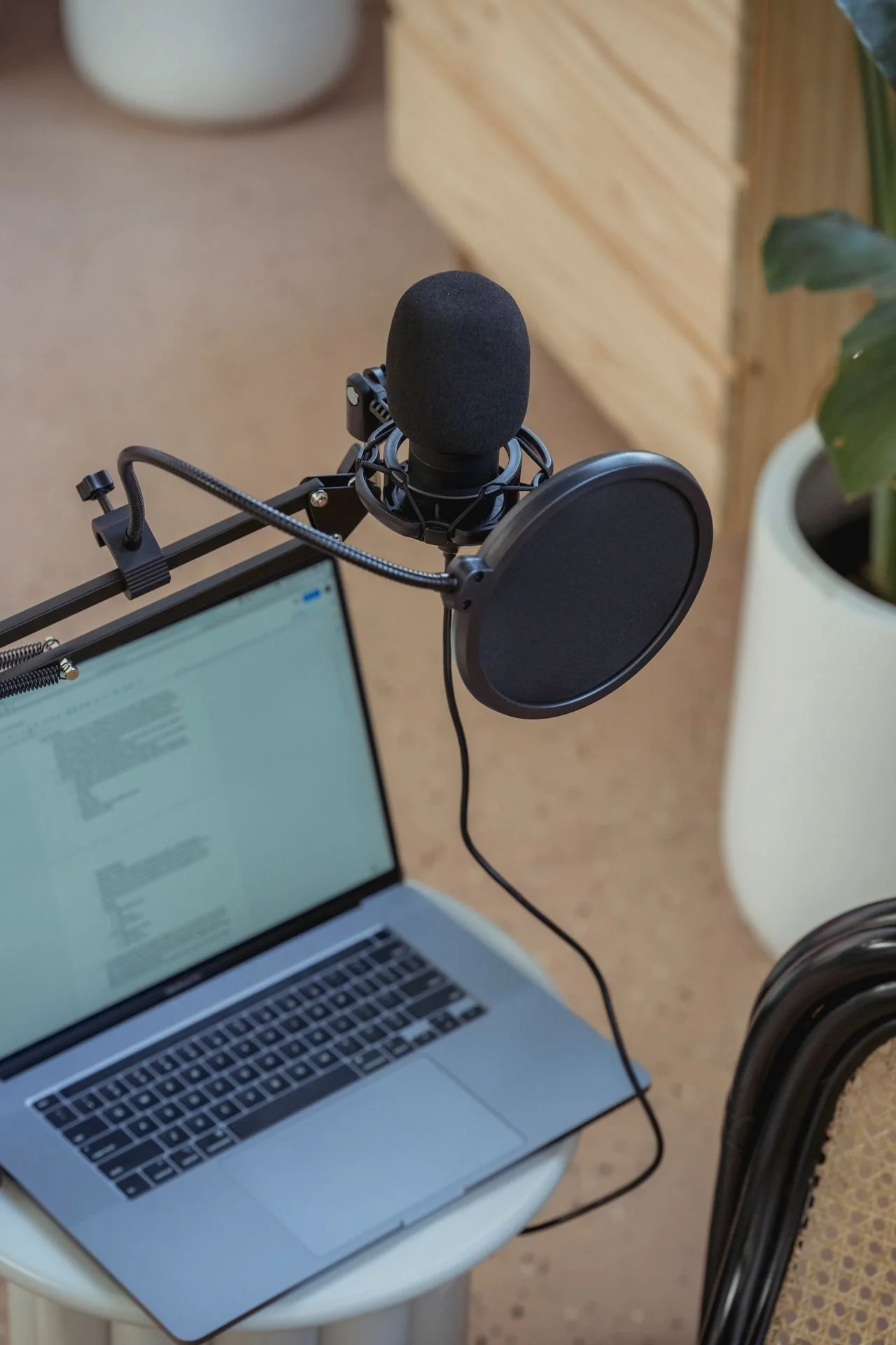 A laptop placed on a small white table with an open document or webpage on the screen, a microphone with a pop filter mounted on a stand, and part of a chair with a black metal frame are visible in the room. A large plant in a white pot and a wooden drawer unit are in the background.