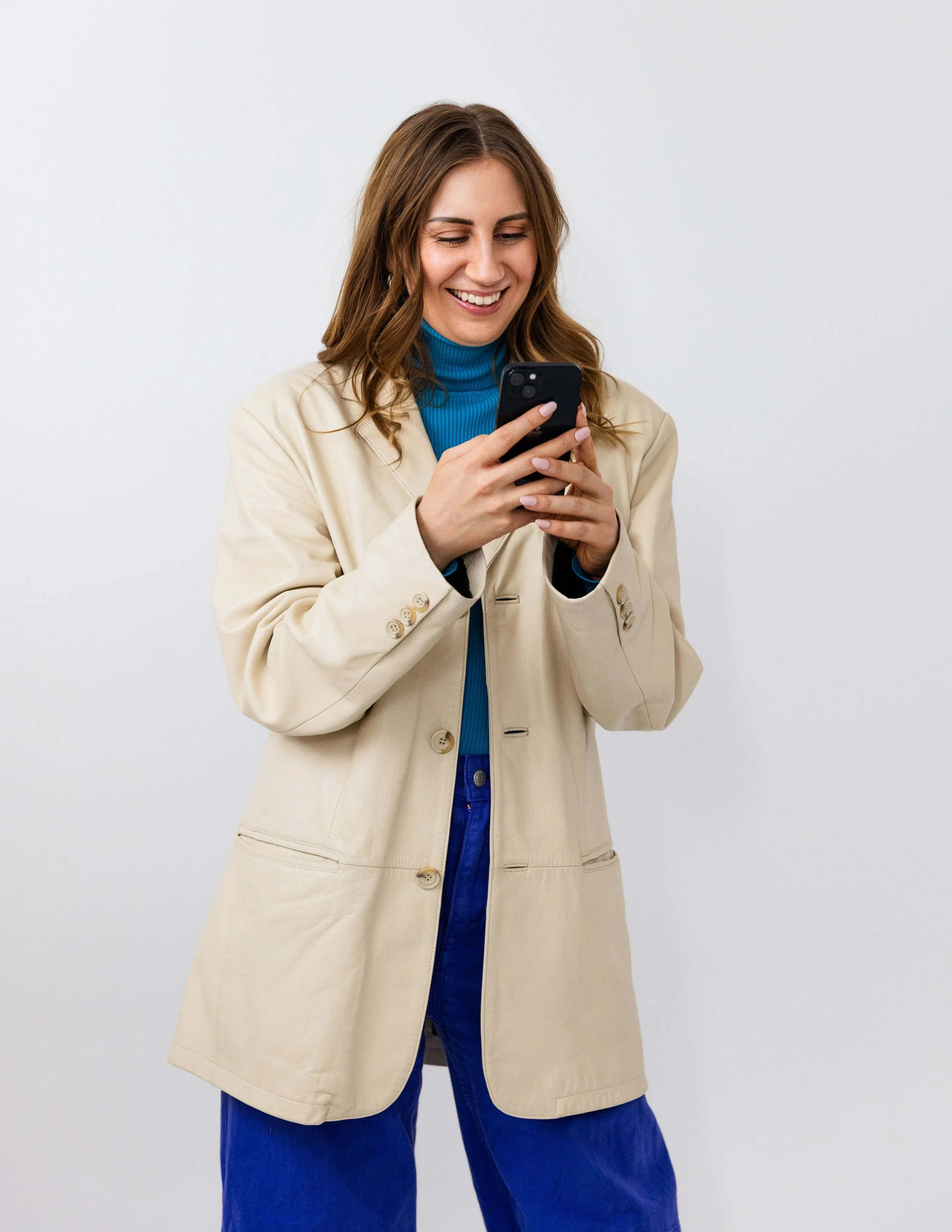 A woman with brown hair wearing a beige coat, blue turtleneck, and blue jeans, smiling while looking at her smartphone.