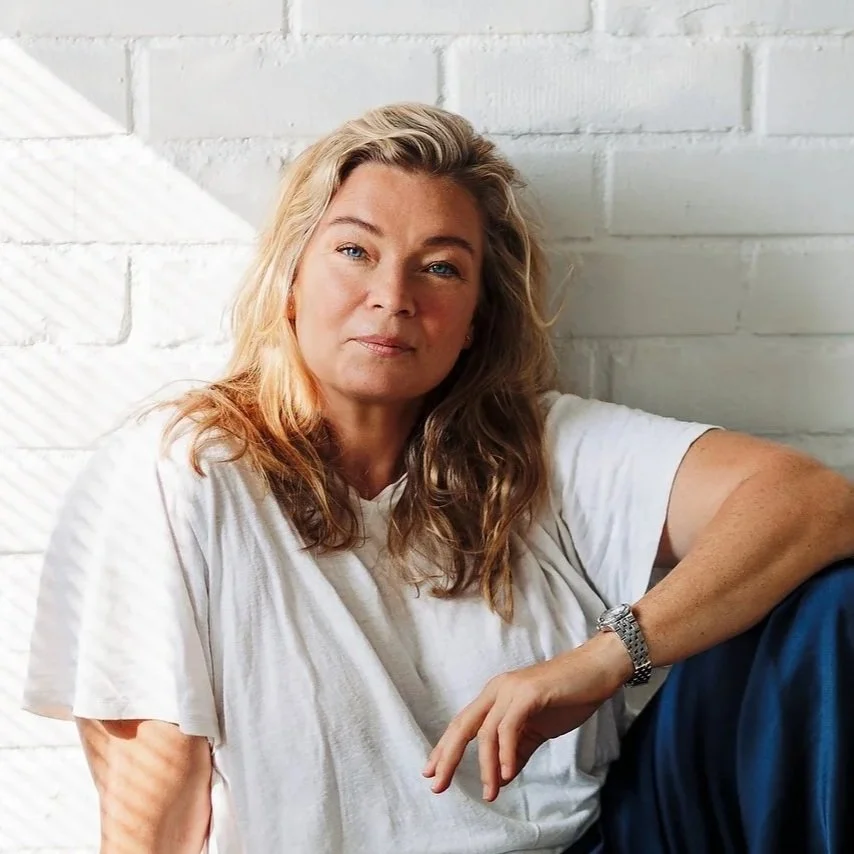 A woman with blonde, wavy hair sitting against a white brick wall wearing a white T-shirt and a silver wristwatch.