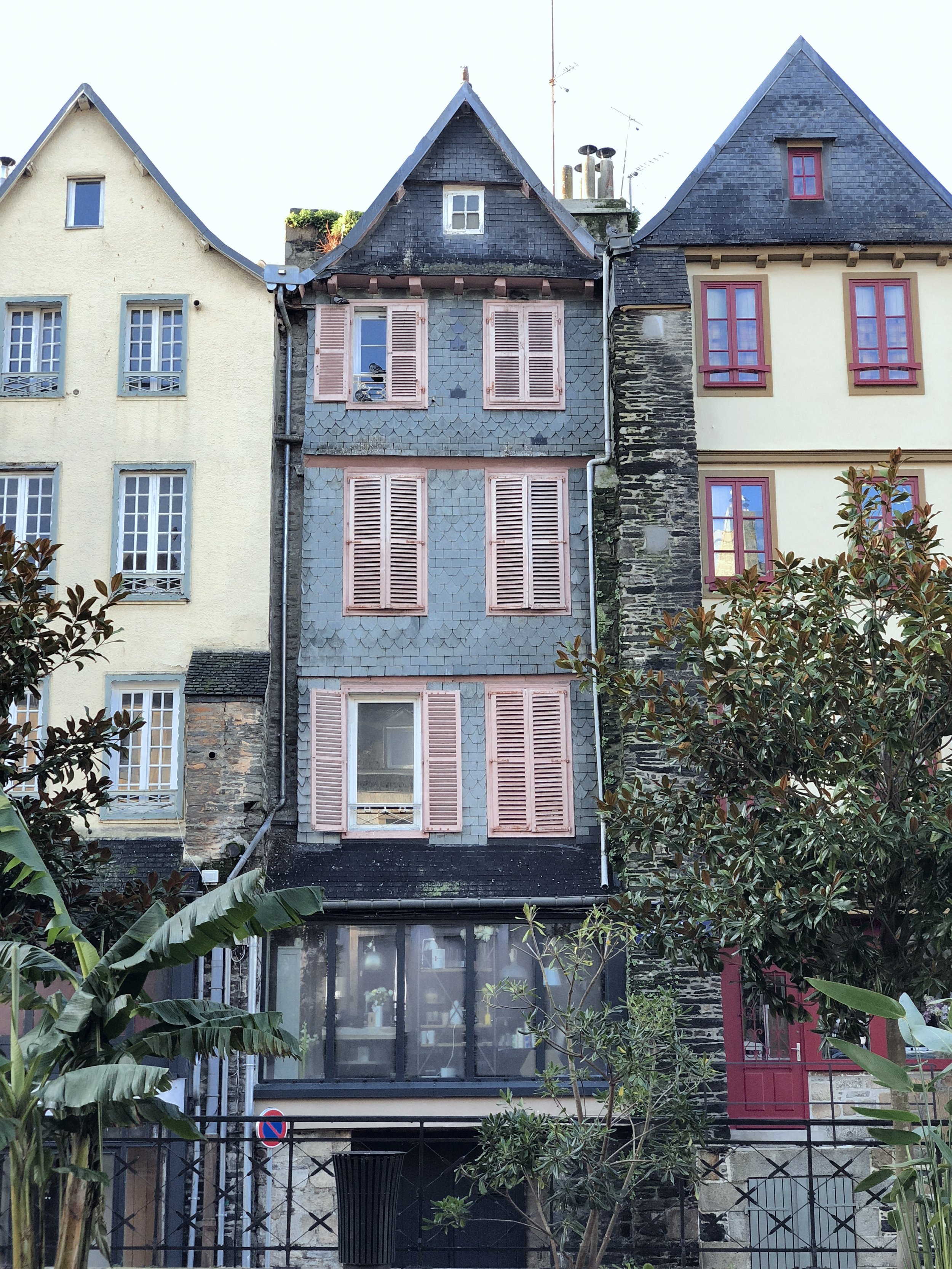 Facade of three old houses with pink-painted wooden shuttered windows, staircase, plants and iron fence.