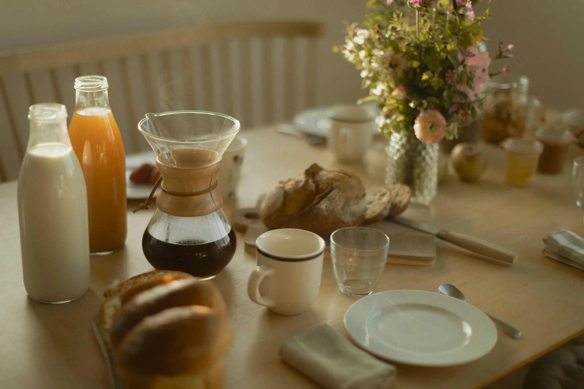 Breakfast table with bottles of milk and juice, a pot of coffee, a plate of bread, a cup, a glass, flowers in a vase, and small jars of jam.