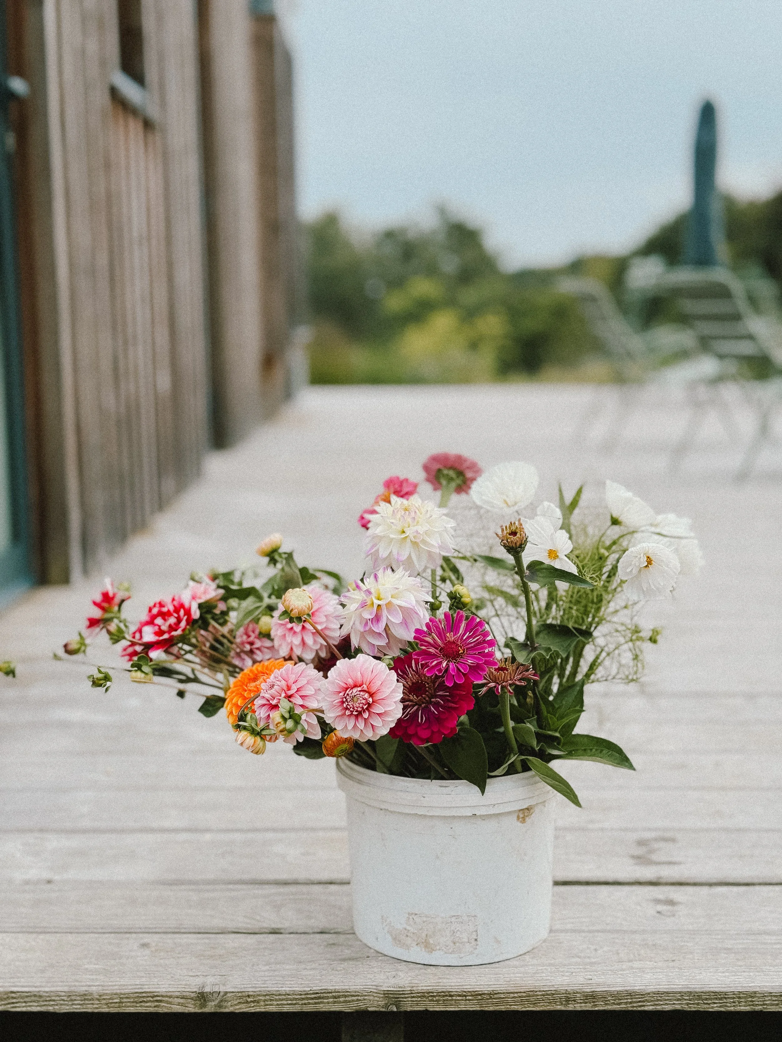 Un bouquet de fleurs colorées dans un pot blanc sur une table en bois sur une terrasse extérieure.