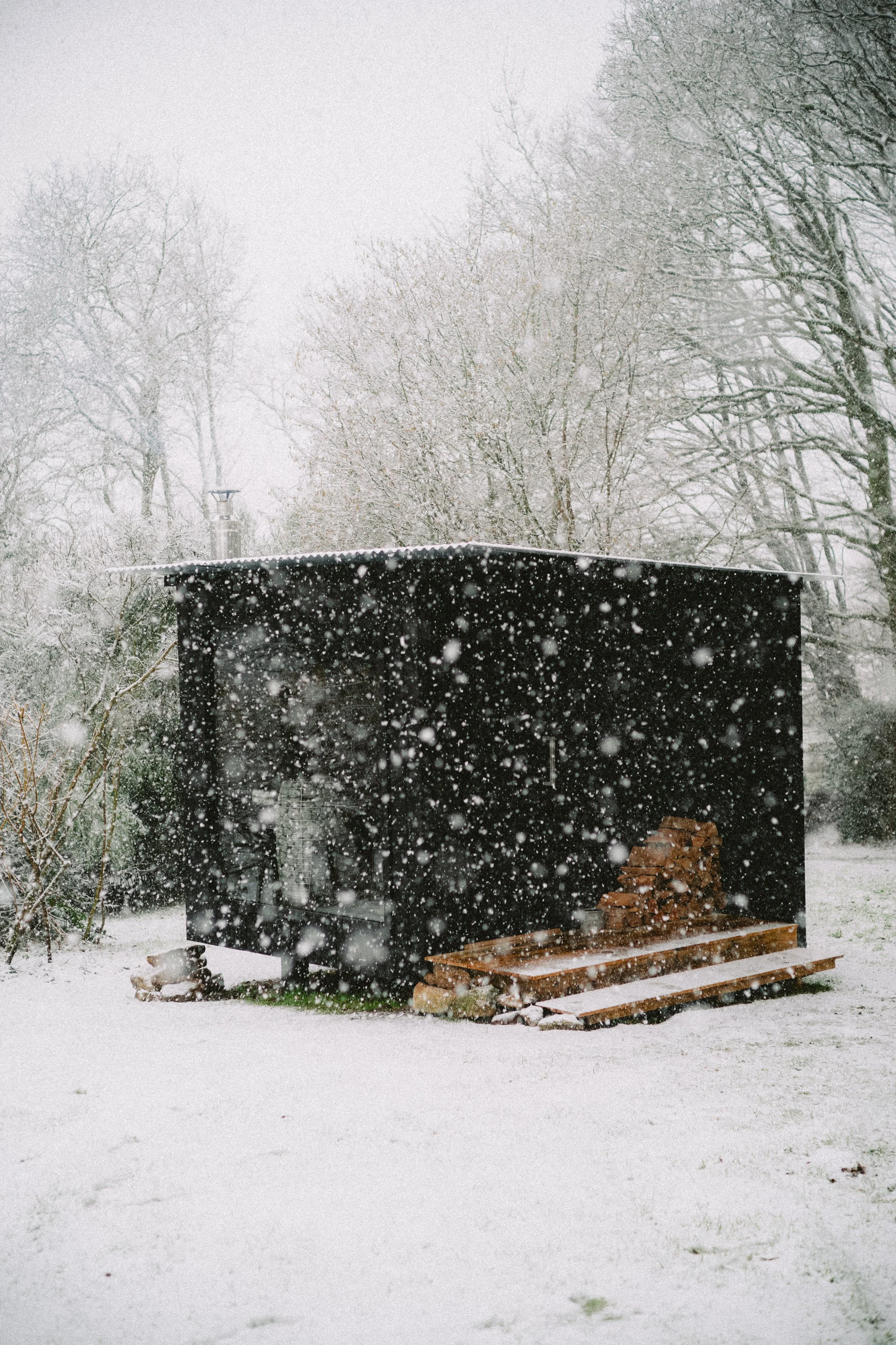 Une cabane noire dans la neige, entourée d'arbres enneigés, avec du bois empilé à l'extérieur.
