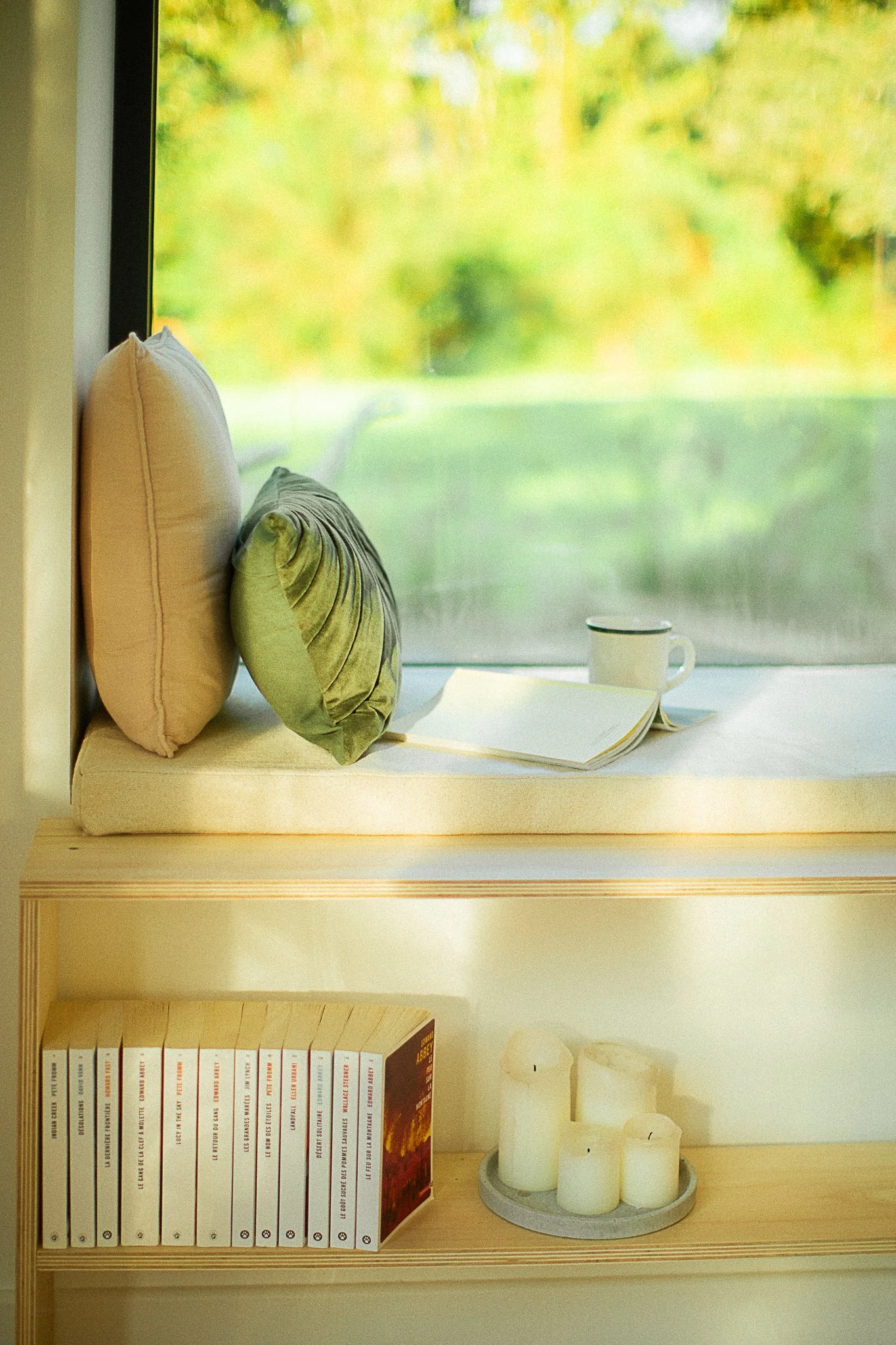 A reading corner near a large window overlooking nature, a cushion, an open book, a white cup, and a row of books with a candle.