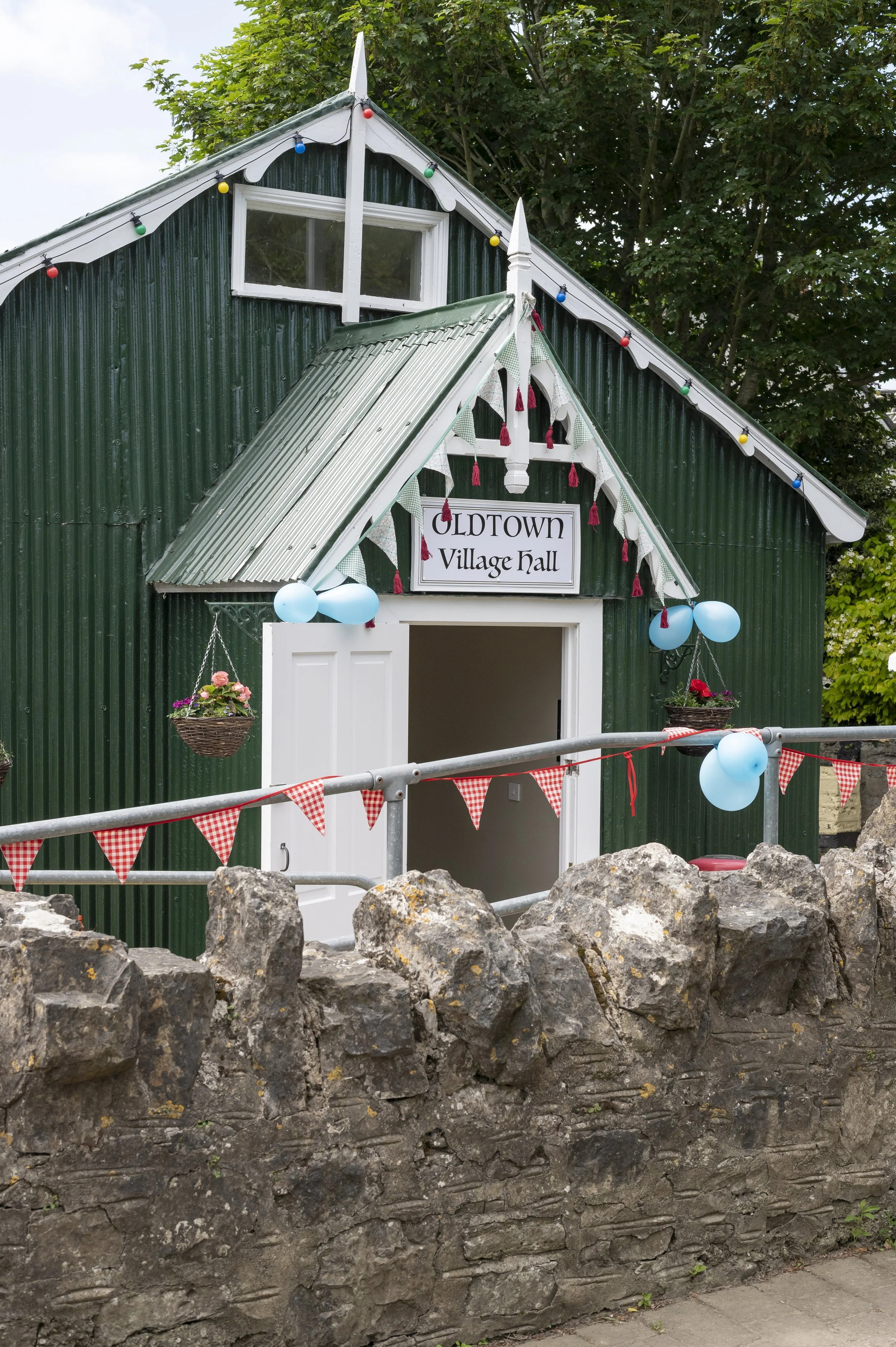 A small, green corrugated metal building with a sign that reads 'Oldtown Village Hall' decorated with string lights, balloons, and flower baskets, set outside with a stone wall in the foreground.