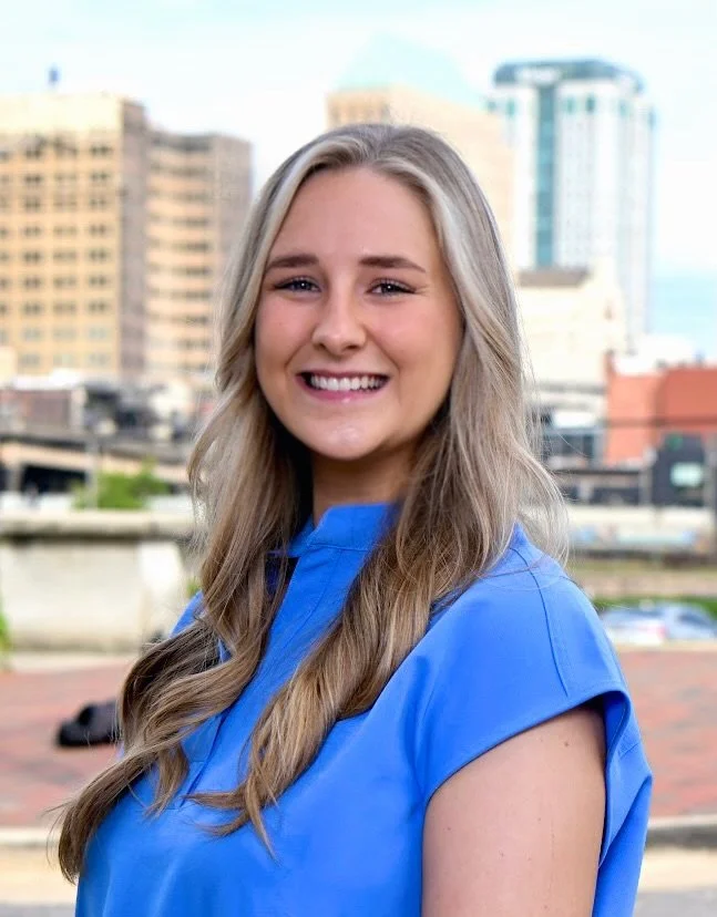 A smiling woman with long blonde hair wearing a blue top, standing outdoors with an urban cityscape background.