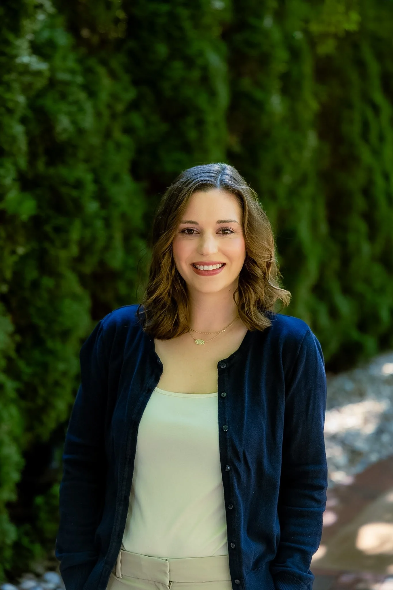 A young woman with shoulder-length wavy brown hair and light skin smiling outdoors, wearing a navy blue button-up shirt over a cream top, standing in front of green foliage.