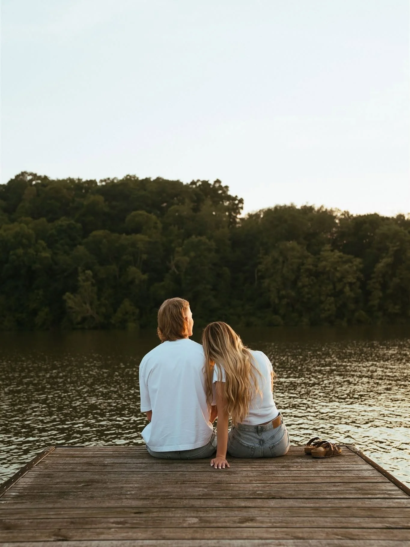 A couple sitting on a wooden dock by a lake, surrounded by trees with a clear sky overhead.
