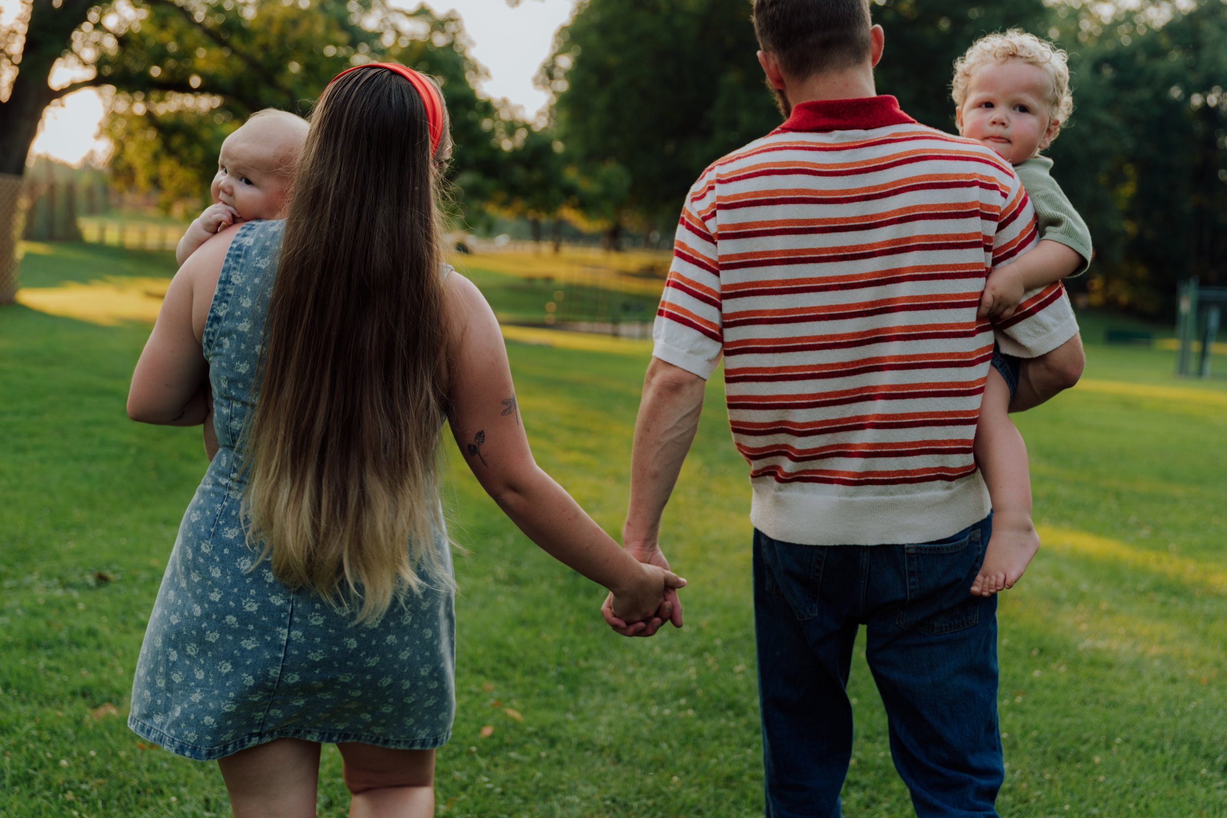 A family of four walking hand in hand in a park during the late afternoon or early evening. The adults are holding hands, with the child with curly blonde hair comfortably resting on the father's shoulder, and the other child with blonde hair and a p