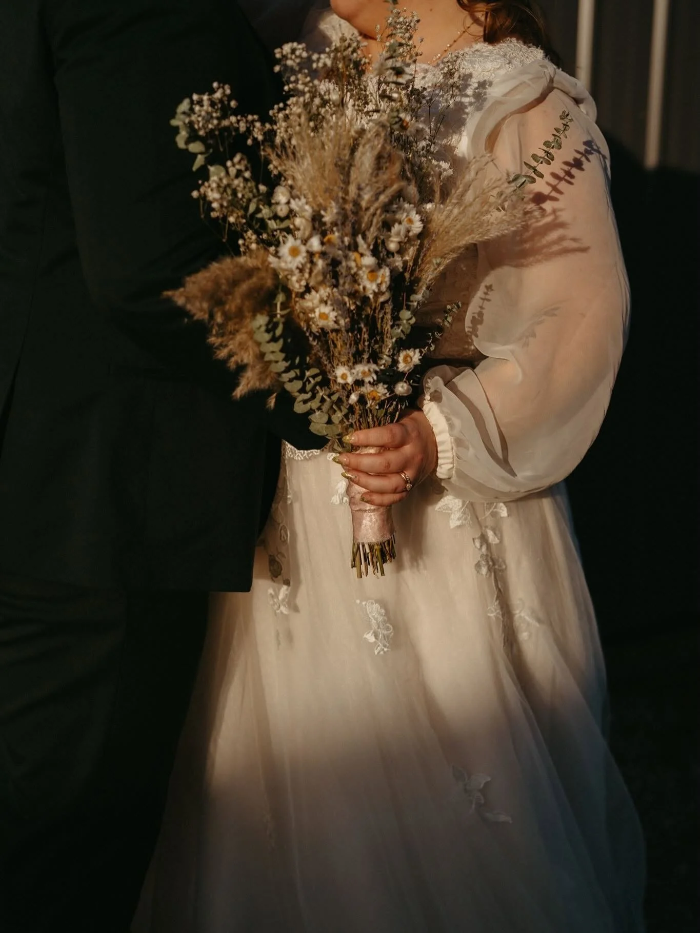 A bride and groom holding a bouquet of dried flowers, including daisies and pampas grass, during their wedding. The bride is wearing a white lace wedding dress, and the groom is dressed in a black suit. The photo is taken indoors with dark background