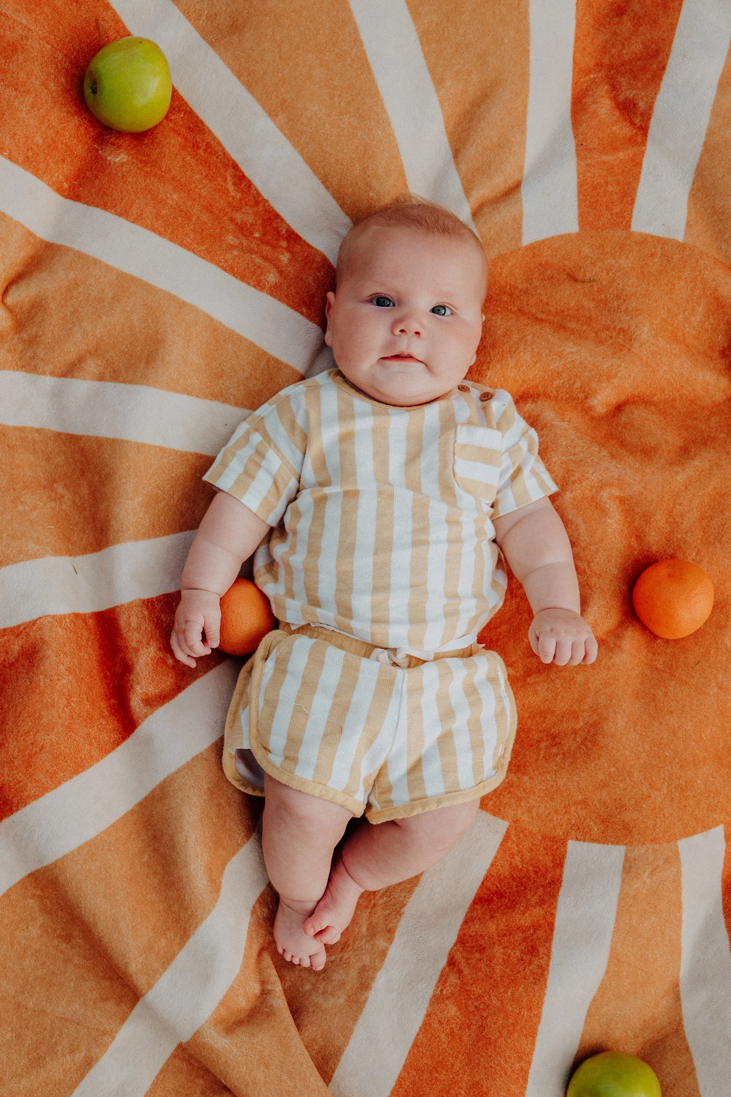 A baby lying on a large orange and white striped towel with a green apple and two oranges nearby.