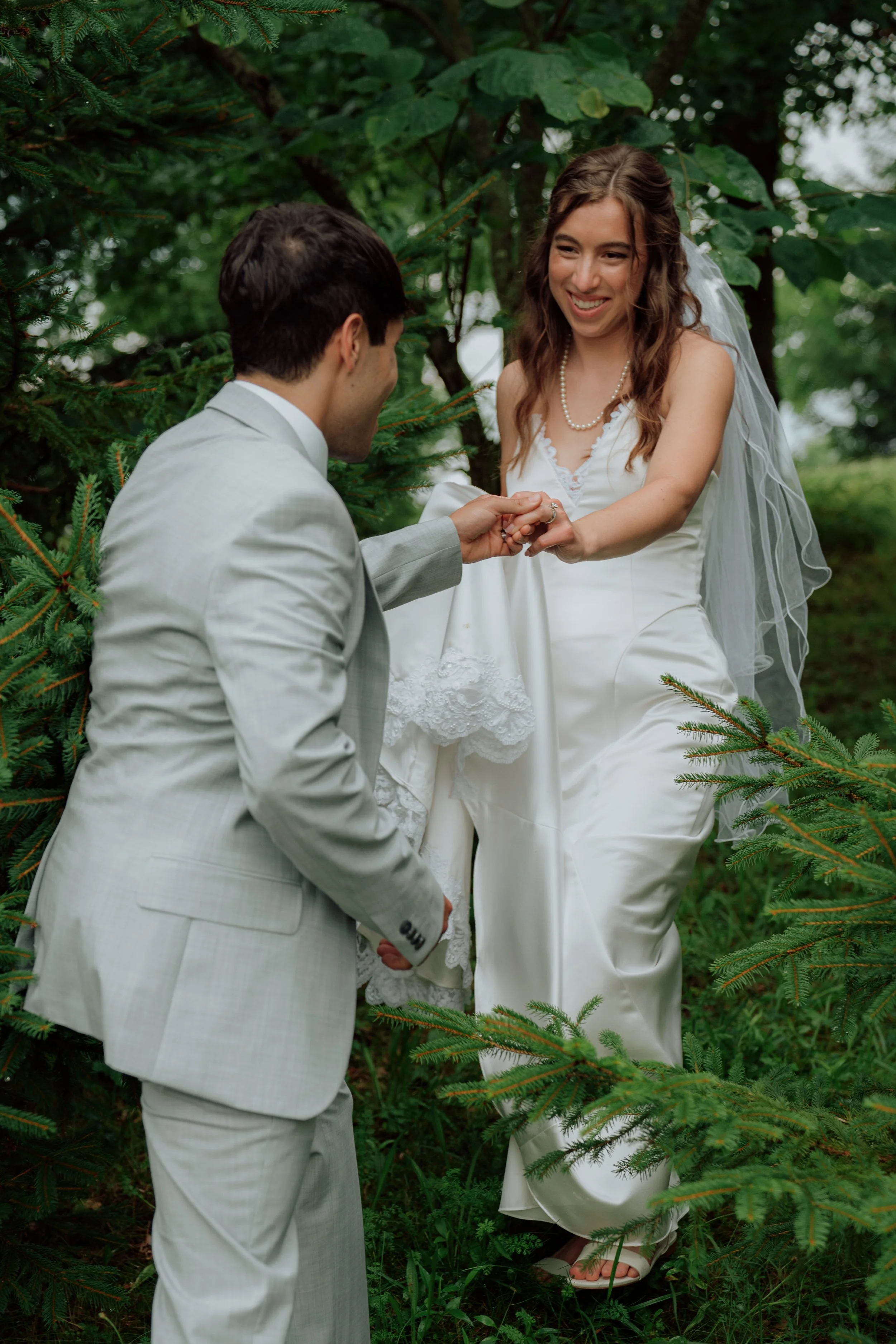 A man in a light suit kneeling and holding hands with a woman in a white dress and veil. They are outdoors among green trees, and the woman is smiling.