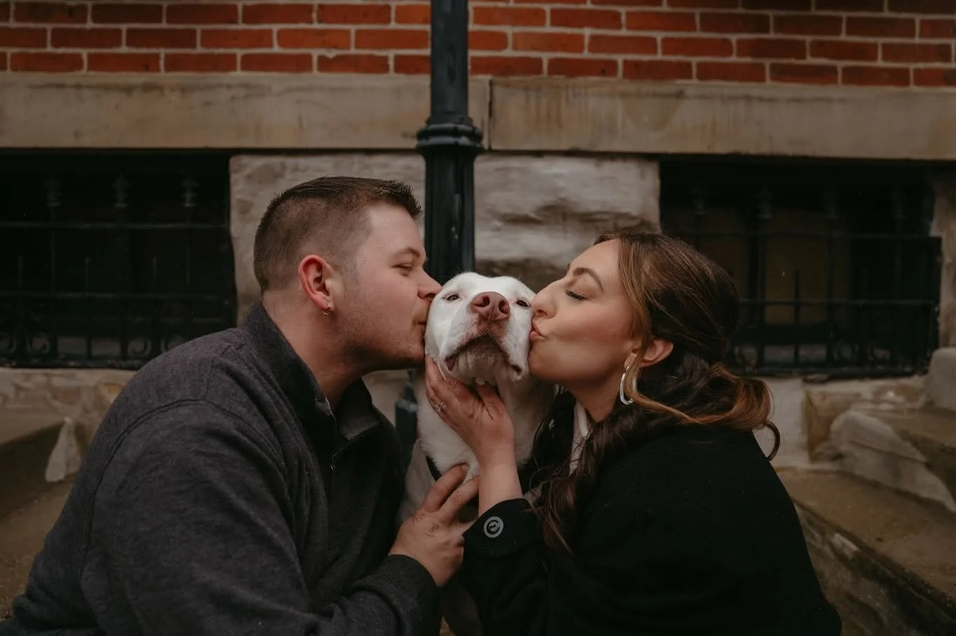 A couple gently kisses their white dog on each cheek in front of a brick fireplace.