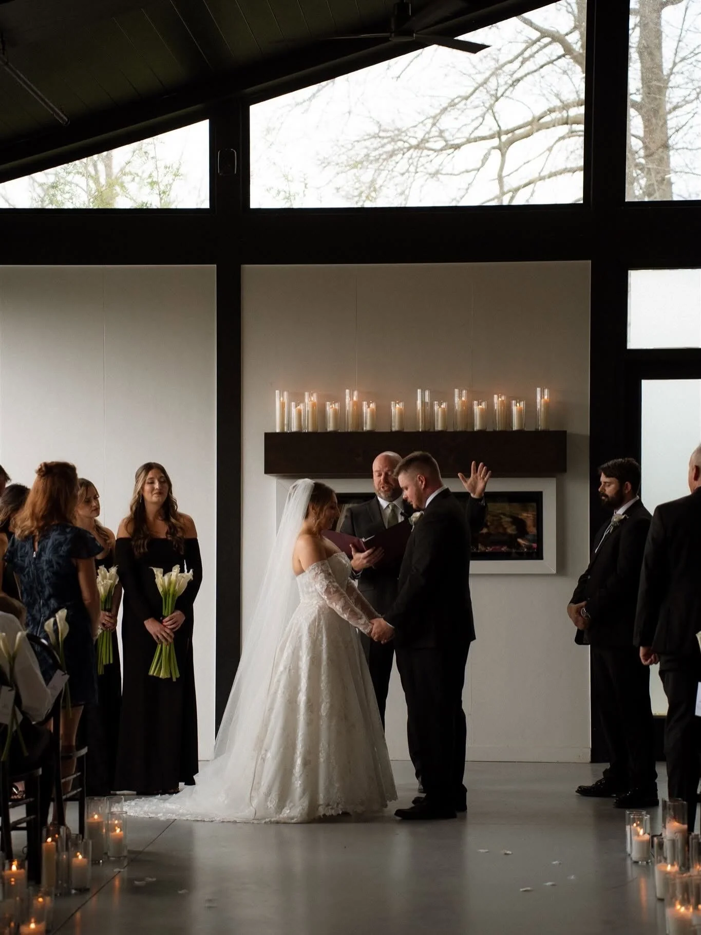 A wedding ceremony with a bride and groom holding hands, surrounded by their wedding party in a modern indoor venue with candles and large windows.