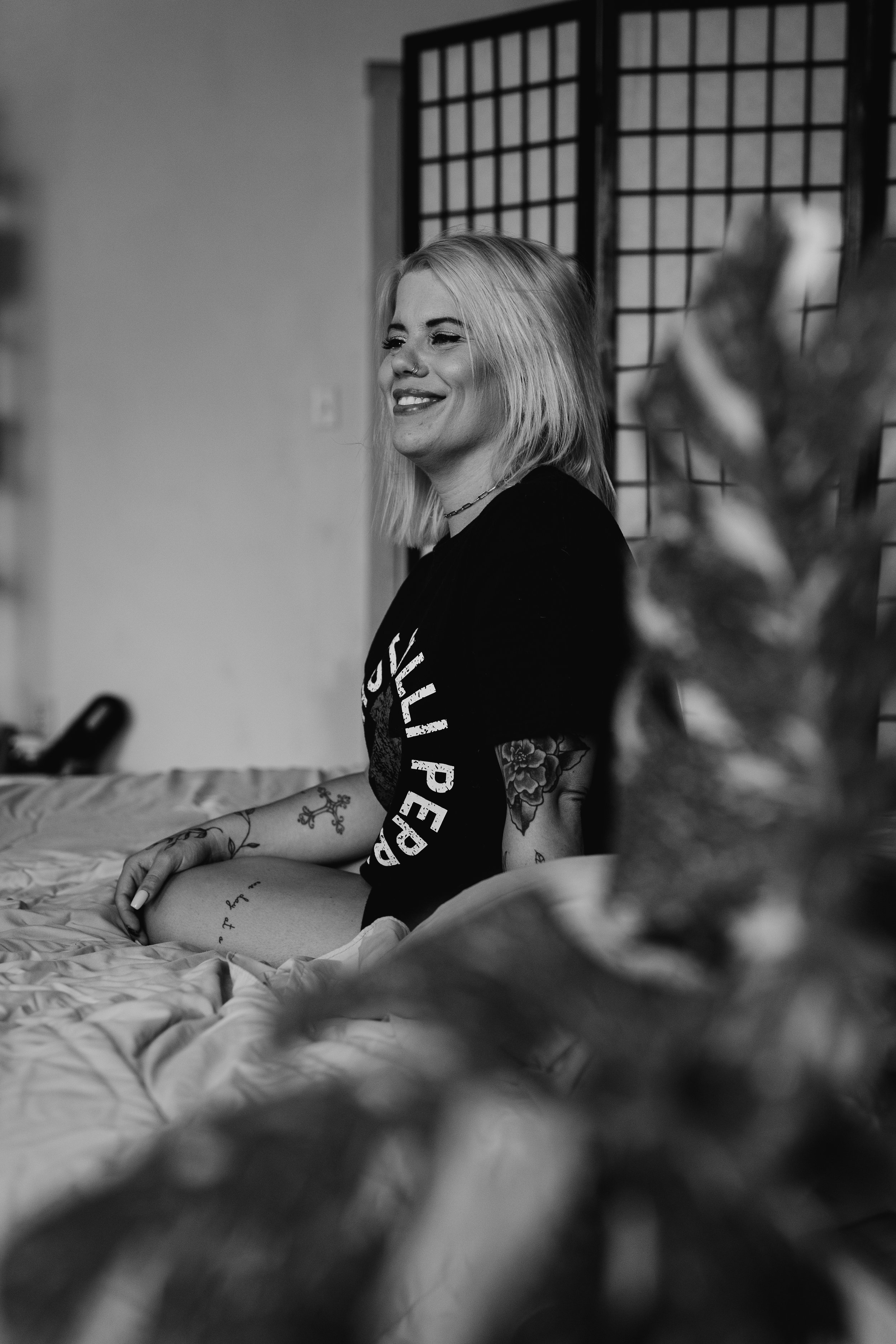 A black and white photo of a smiling woman with shoulder-length blonde hair, tattoos on her arm and leg, sitting on a bed, with a background that includes a folding room divider with a grid pattern.