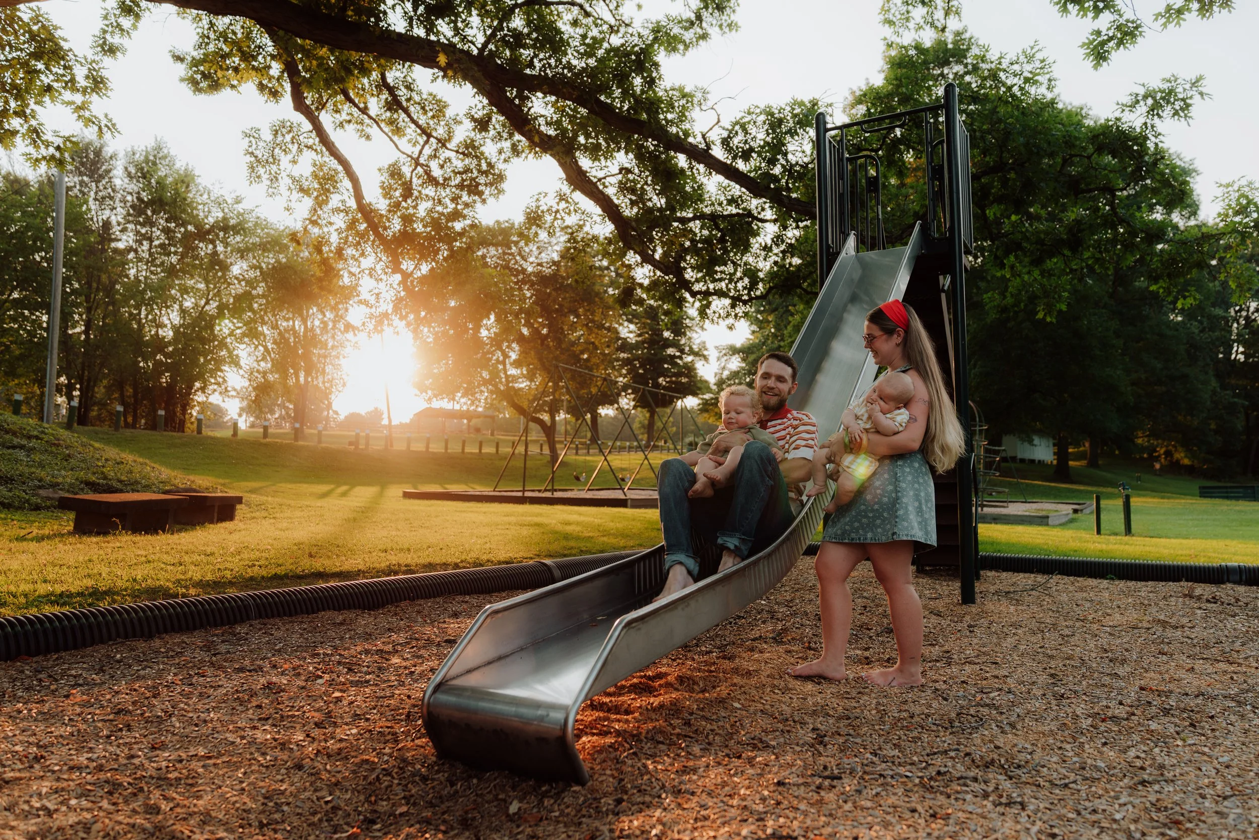 A family of four enjoying a sunset at a playground; mother, father, and two young children, with the slide and trees in the background.