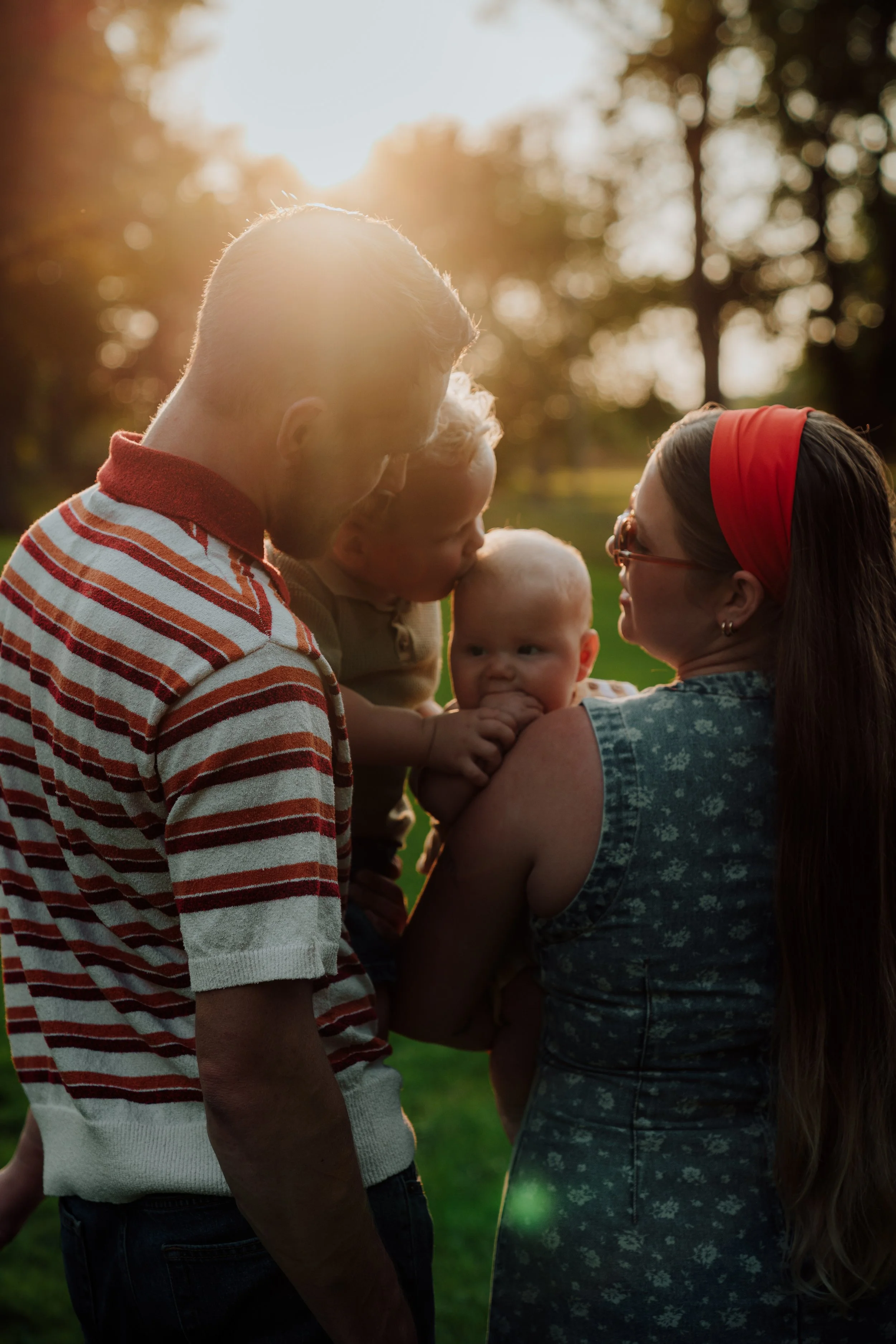 A family of four (a man, a woman, and two children) standing outdoors at sunset, with trees in the background, engaging in an intimate moment of affection and connection.