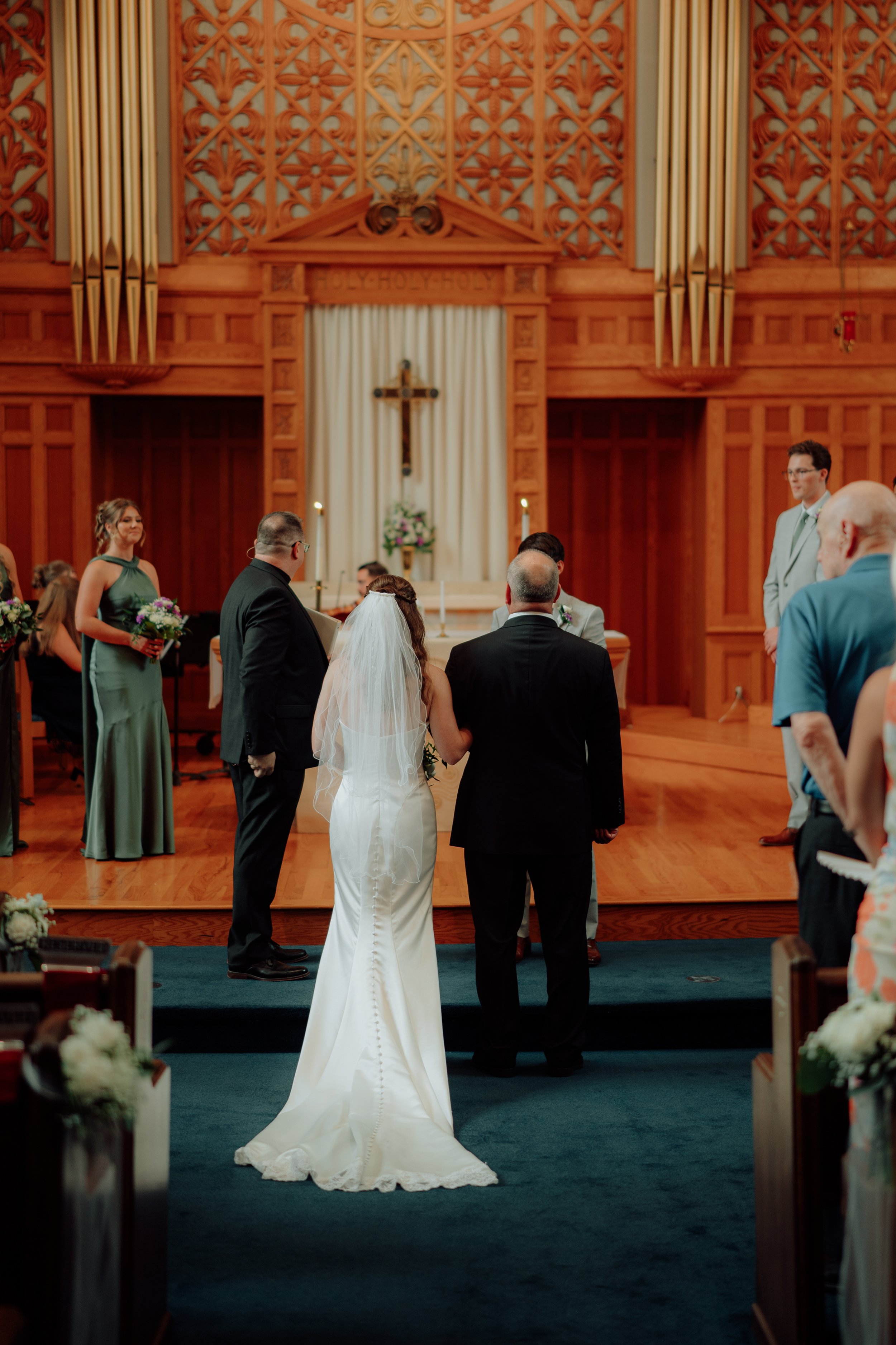 A bride in a white wedding dress with a veil standing at the altar during a wedding ceremony in a church. The groom and guests are present, with church wooden interior and religious symbols in the background.