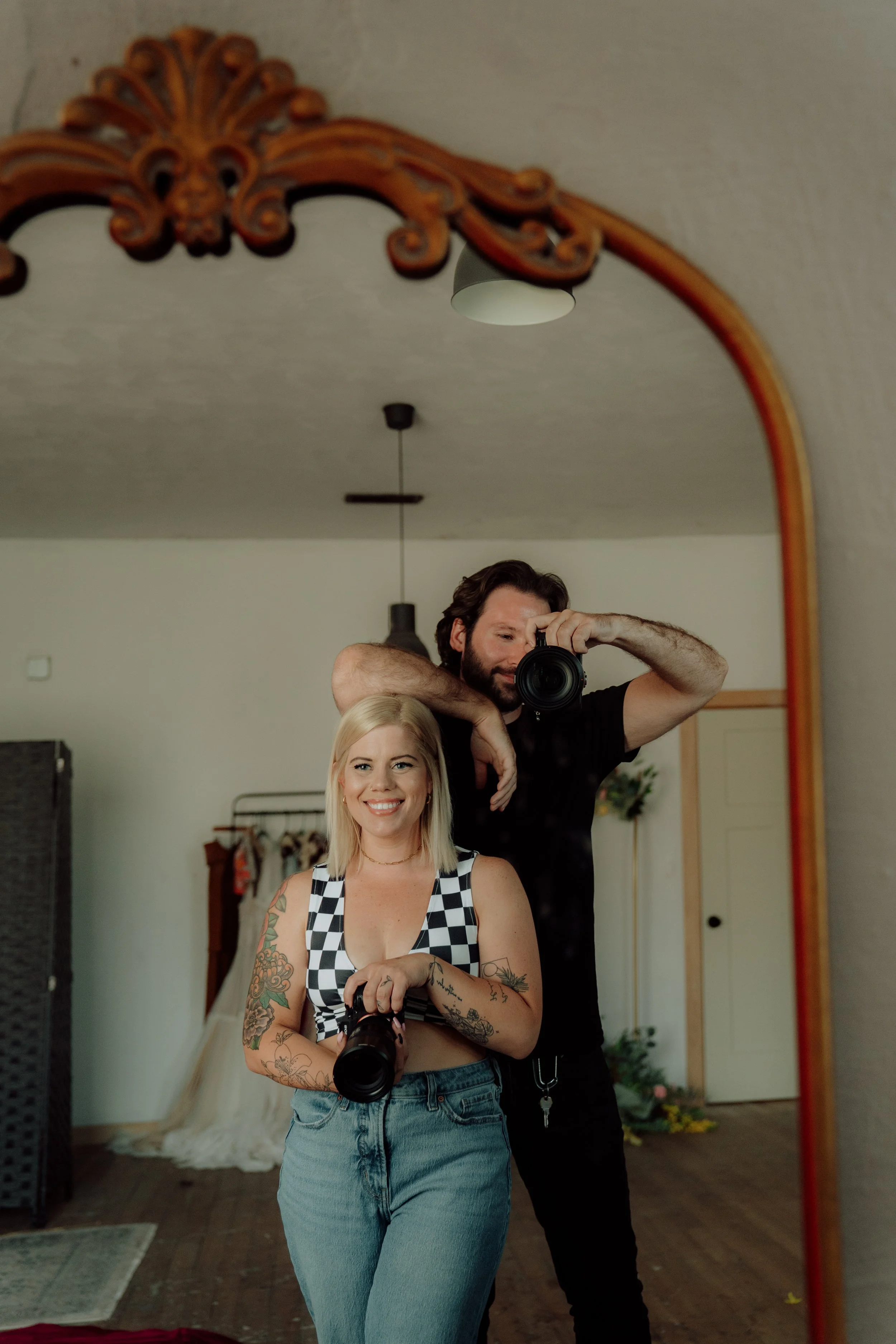 A woman with blonde hair and tattoos on her arms holding a camera, standing in front of a mirror with a man taking a photo of her.