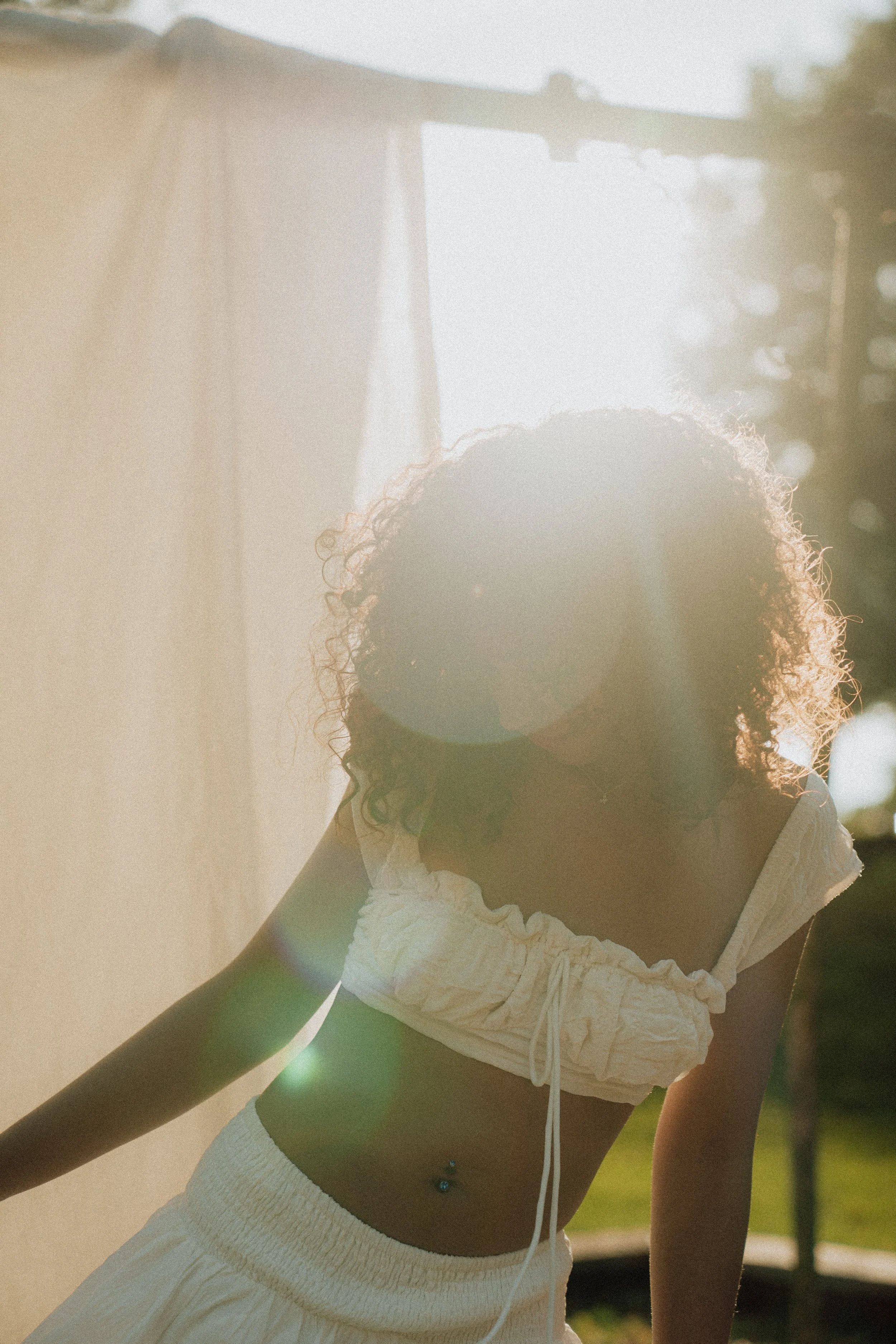 A woman with curly hair in a white top, wearing white shorts and a belly button piercing, standing outdoors in sunlight, with a blurred background of trees and a building.