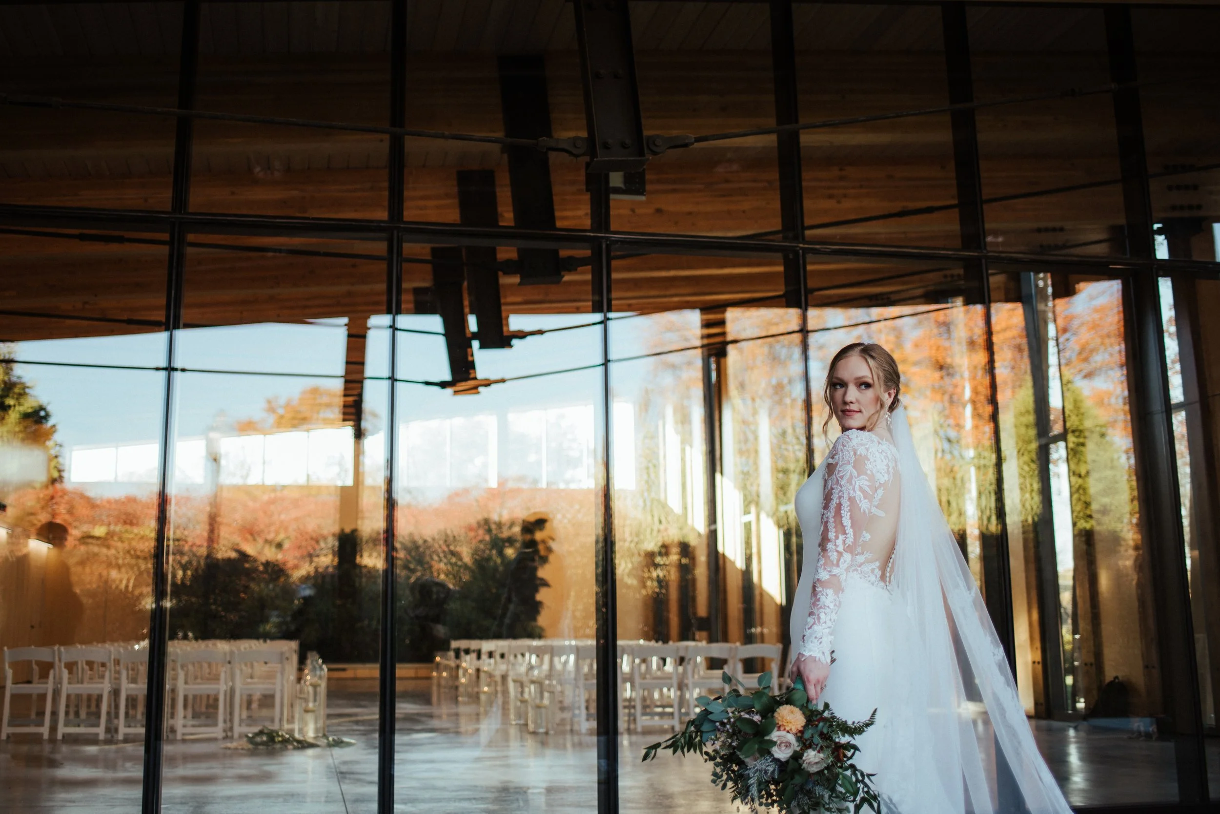 A bride dressed in white lace wedding gown standing inside a glass-walled venue with wooden ceiling, holding a bouquet with greenery and flowers. Outside autumn trees are visible.