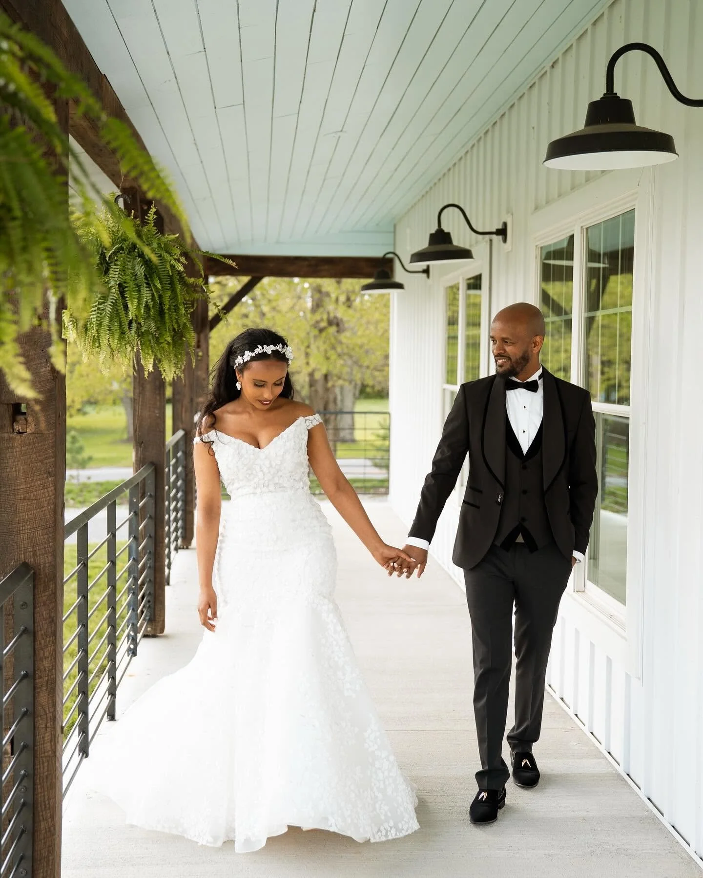 Bride and groom holding hands walking on porch, bride in white wedding dress with floral headband, groom in black tuxedo, outdoor setting with trees.