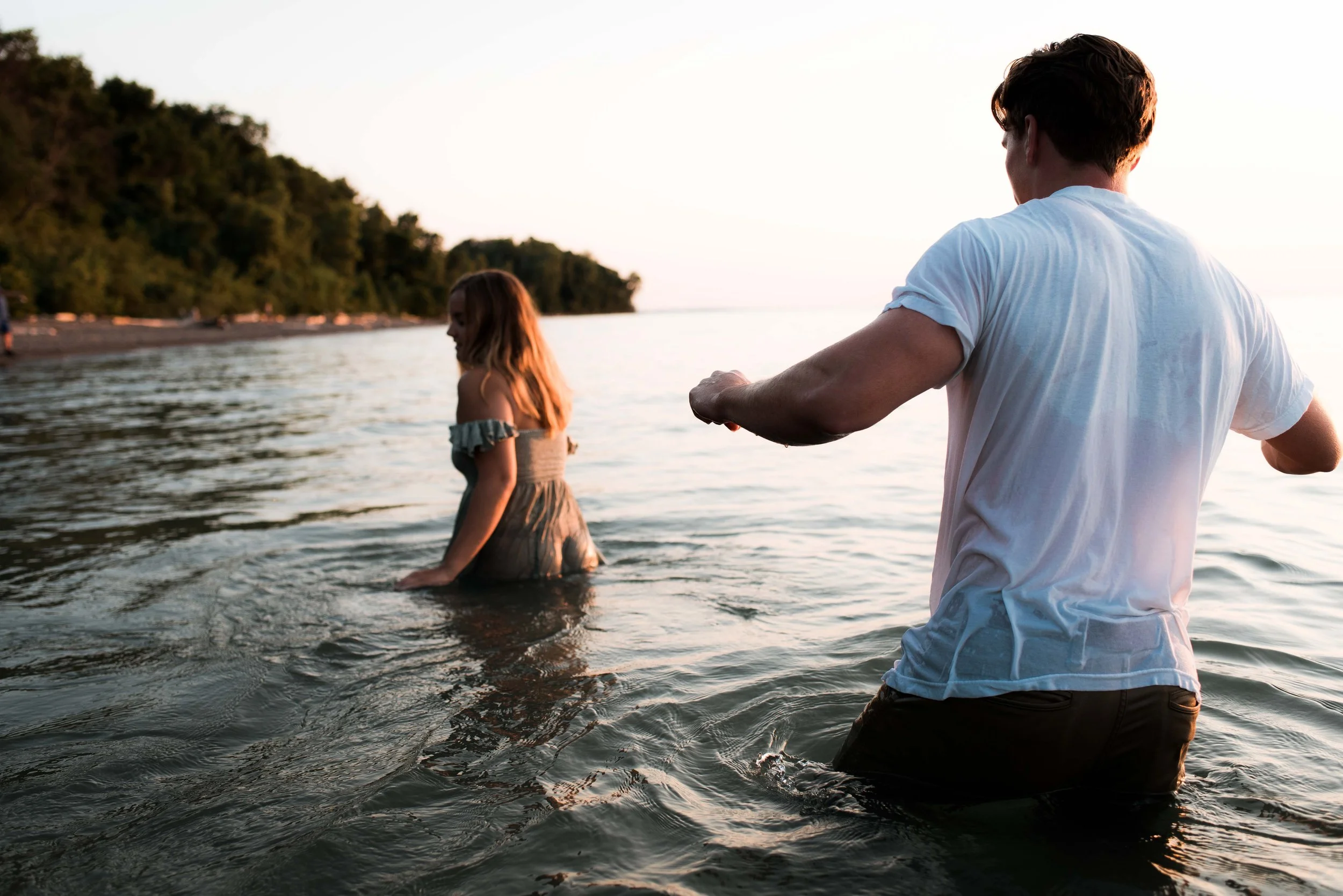 A man in a white T-shirt and black shorts wading in the water while a woman in a dress sits in the water near the shoreline of a beach at sunset, with a tree-lined hill in the background.