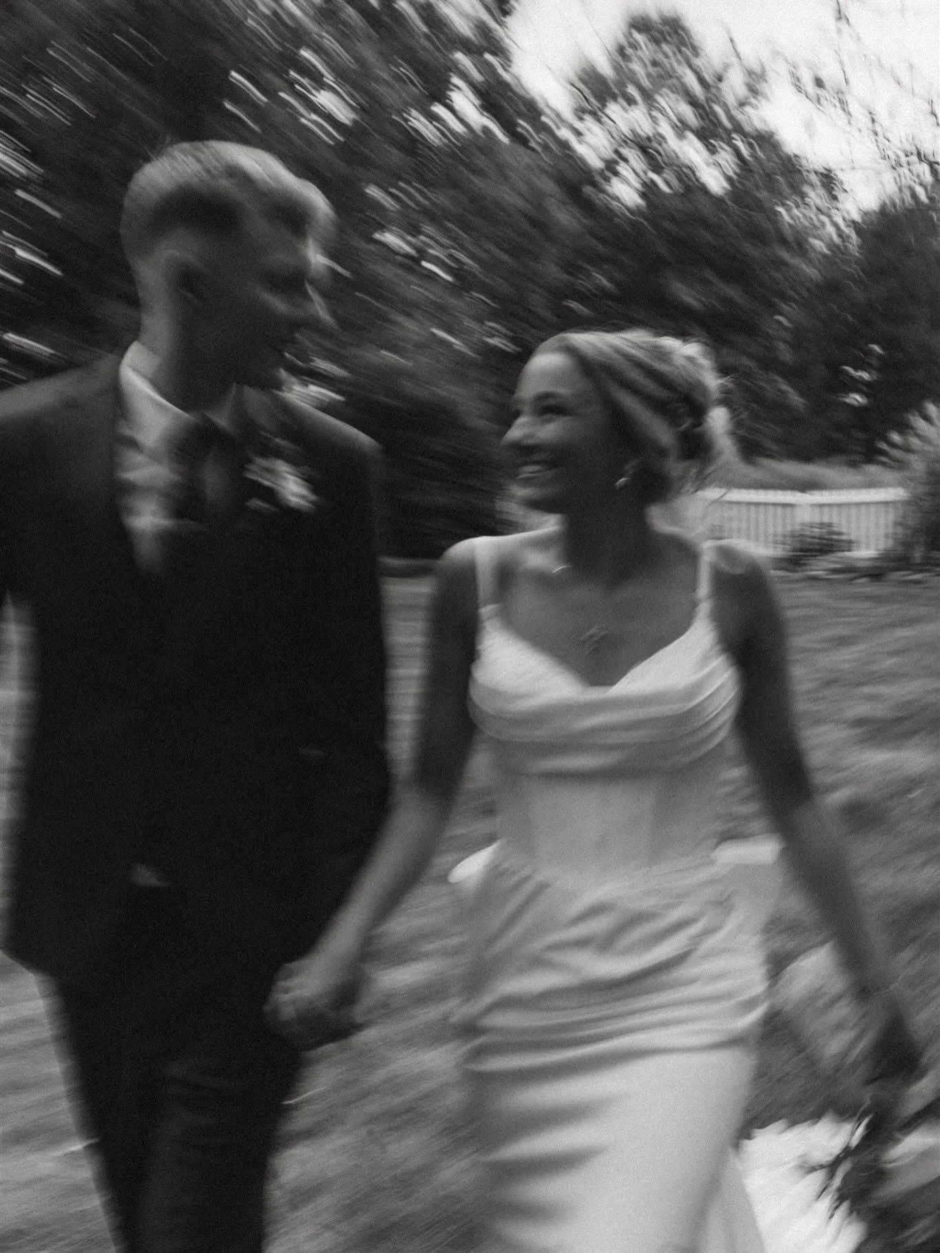 A black and white photo of a man and woman smiling and walking outdoors, holding hands. The man is dressed in a suit with a boutonniere, and the woman is wearing a wedding dress with a flower in her hair. They appear happy and are moving, with a blur
