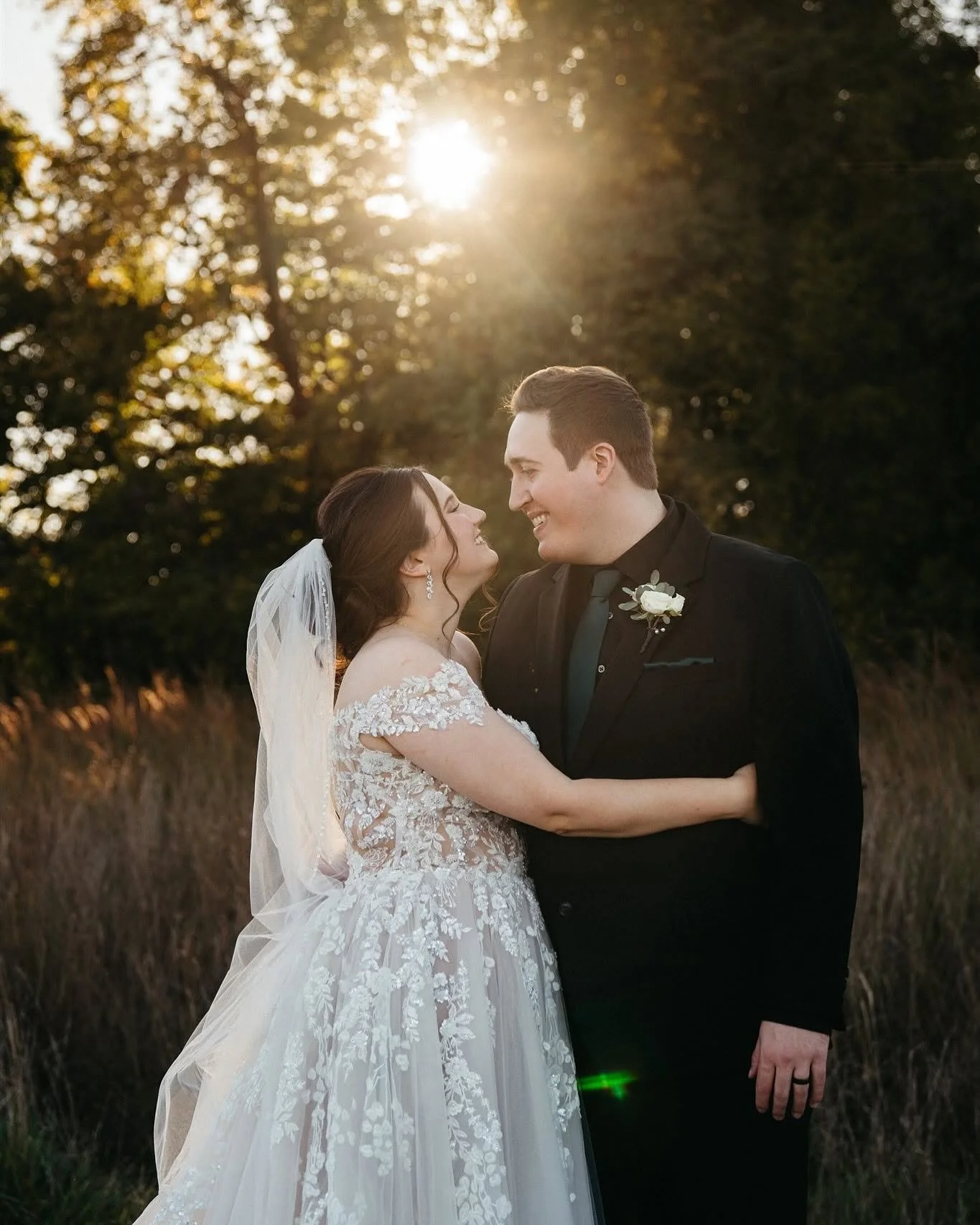 A bride and groom are standing close together, gazing into each other's eyes and smiling. The bride is wearing a lace wedding gown with off-the-shoulder sleeves and a veil, while the groom is dressed in a black tuxedo with a boutonniere. The background features trees with sunlight streaming through the leaves, creating a warm, romantic atmosphere.