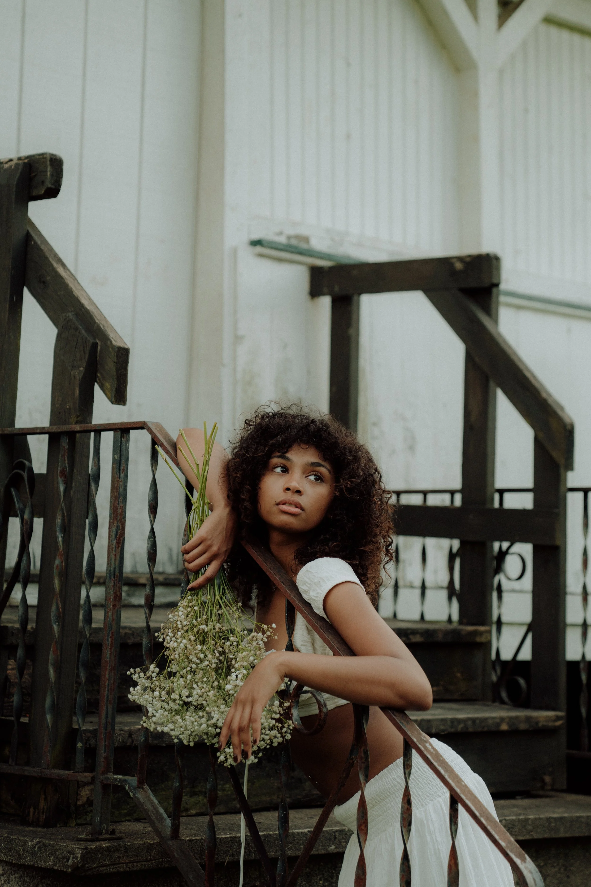 A young woman with curly hair sitting on outdoor stairs, holding a bouquet of white flowers, wearing a white dress, looking to the side with a thoughtful expression.