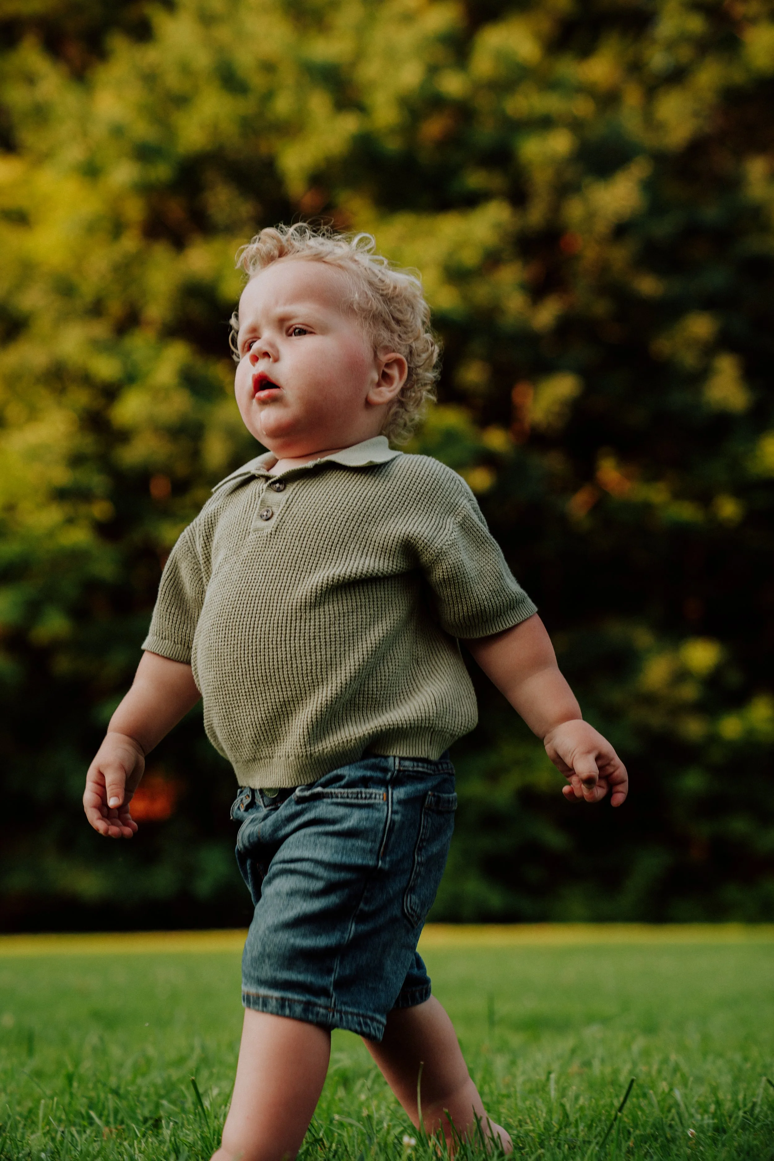 A young boy with curly blonde hair wearing a green polo shirt and denim shorts walking on a grassy field with trees in the background.