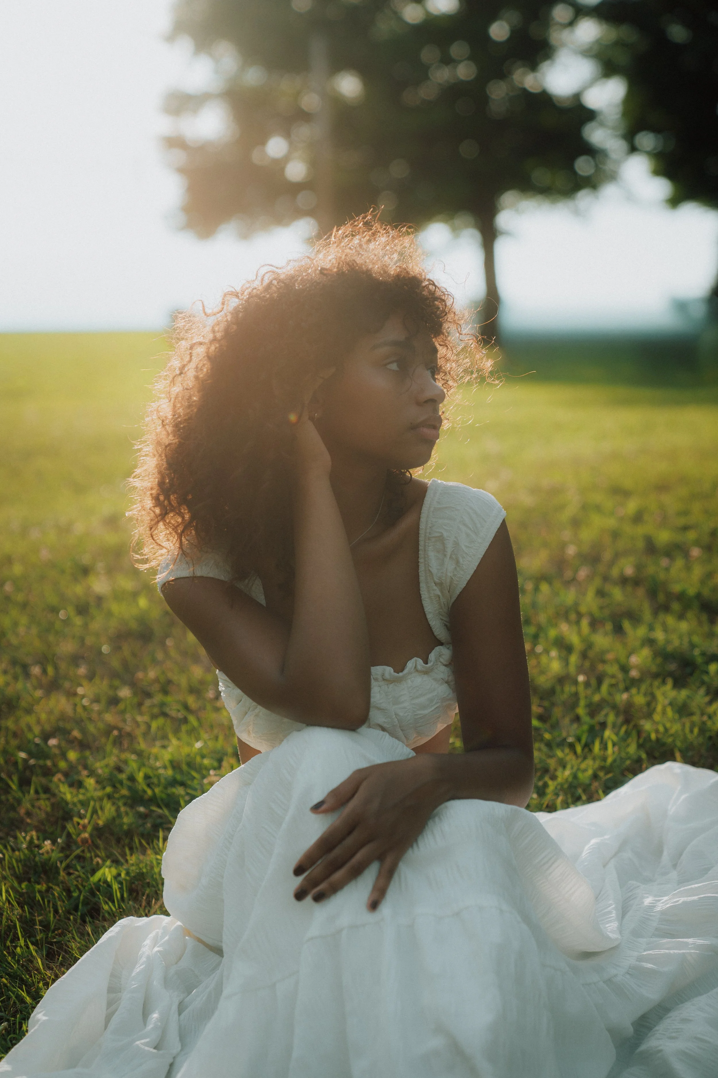 Young woman with curly hair sitting on grass in a white dress, illuminated by sunlight, with trees in the background.