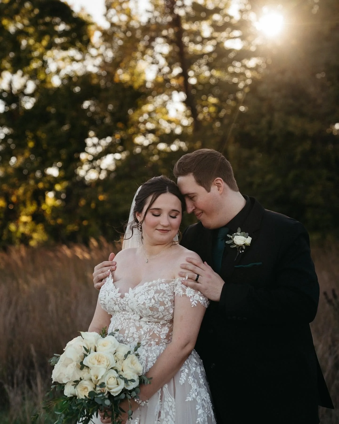 A bride and groom embrace outdoors at sunset, with trees and golden light in the background. The bride is holding a bouquet of white roses and has her eyes closed, while the groom gently touches her shoulders and has his forehead pressed against hers