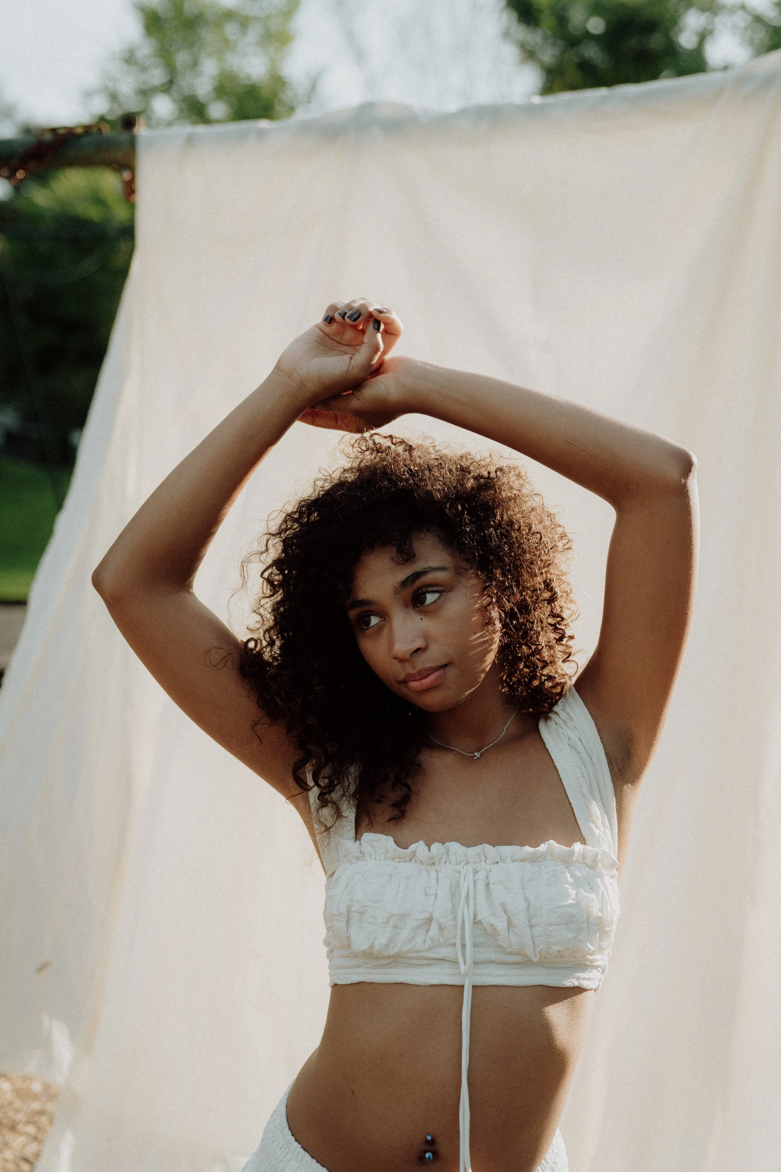 A woman with curly hair stretching her arms above her head outdoors in front of a white fabric backdrop.