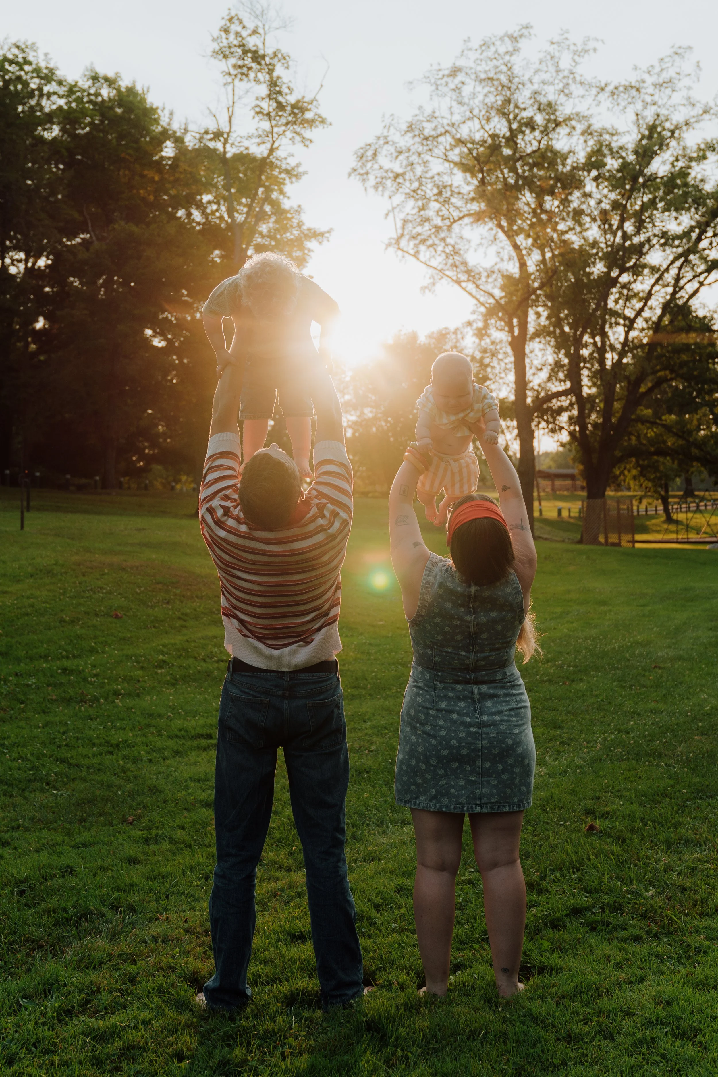 A family of four in a park, two adults lifting children into the air, with trees and a fence in the background during sunset.