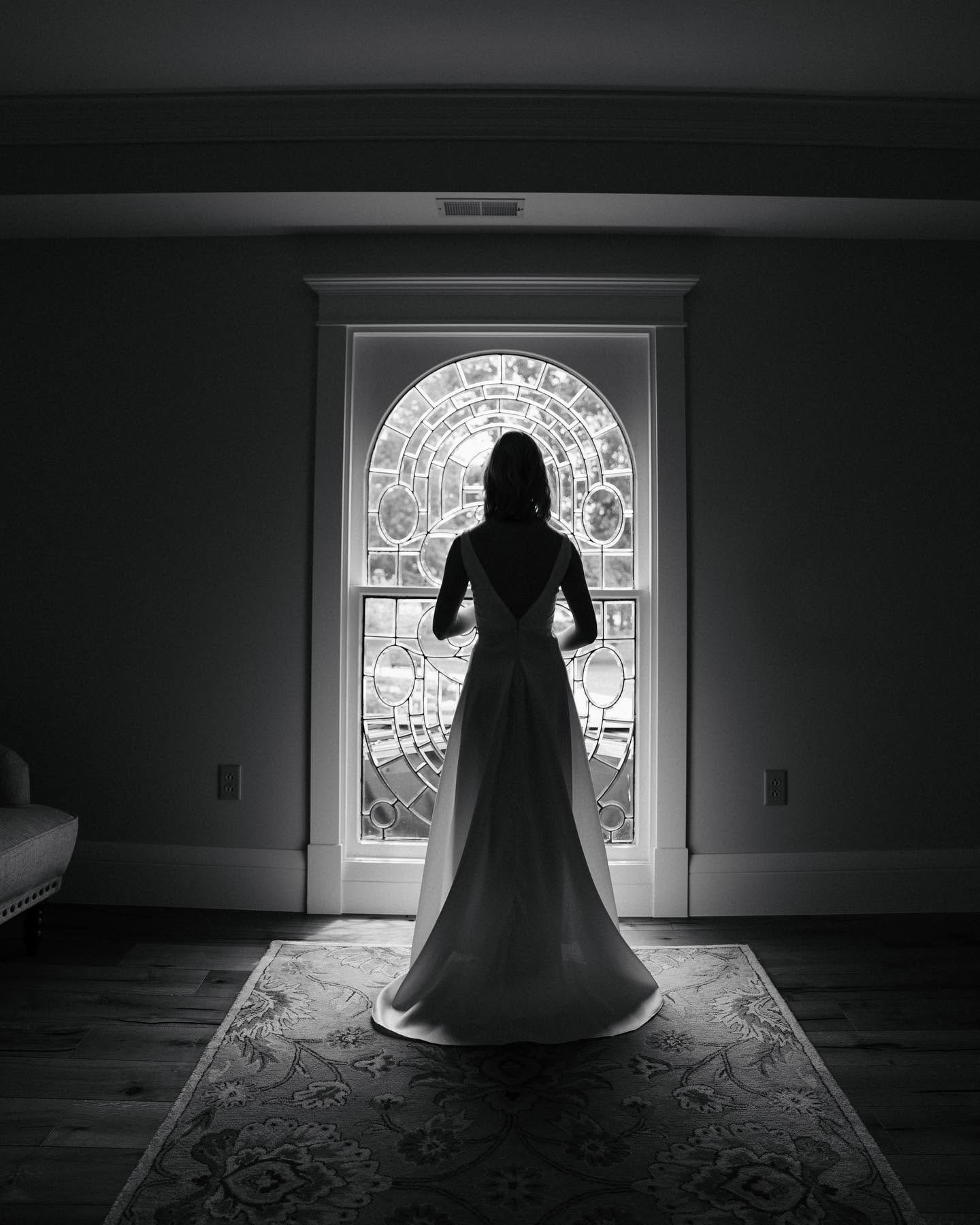 A woman in a wedding dress standing indoors in front of a decorative stained glass window, silhouetted against the light.