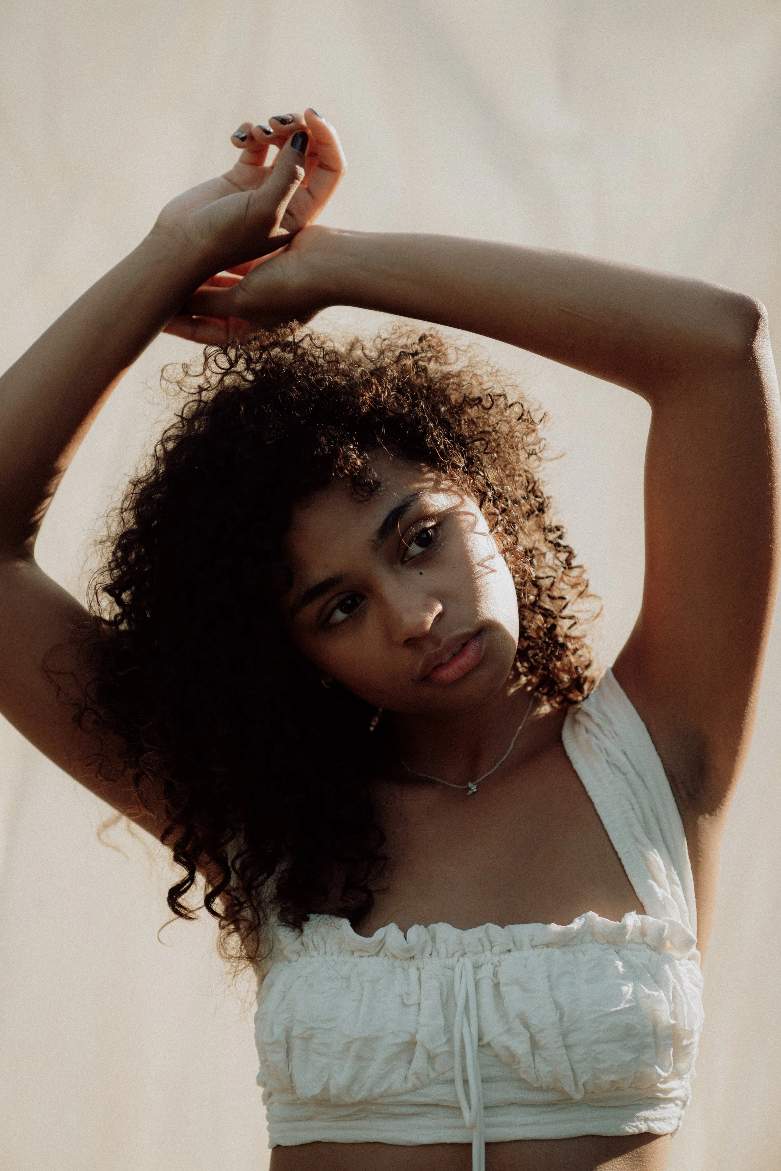 A young woman with curly hair wearing a white sleeveless top and shorts, posing with arms raised above her head against a plain light background.