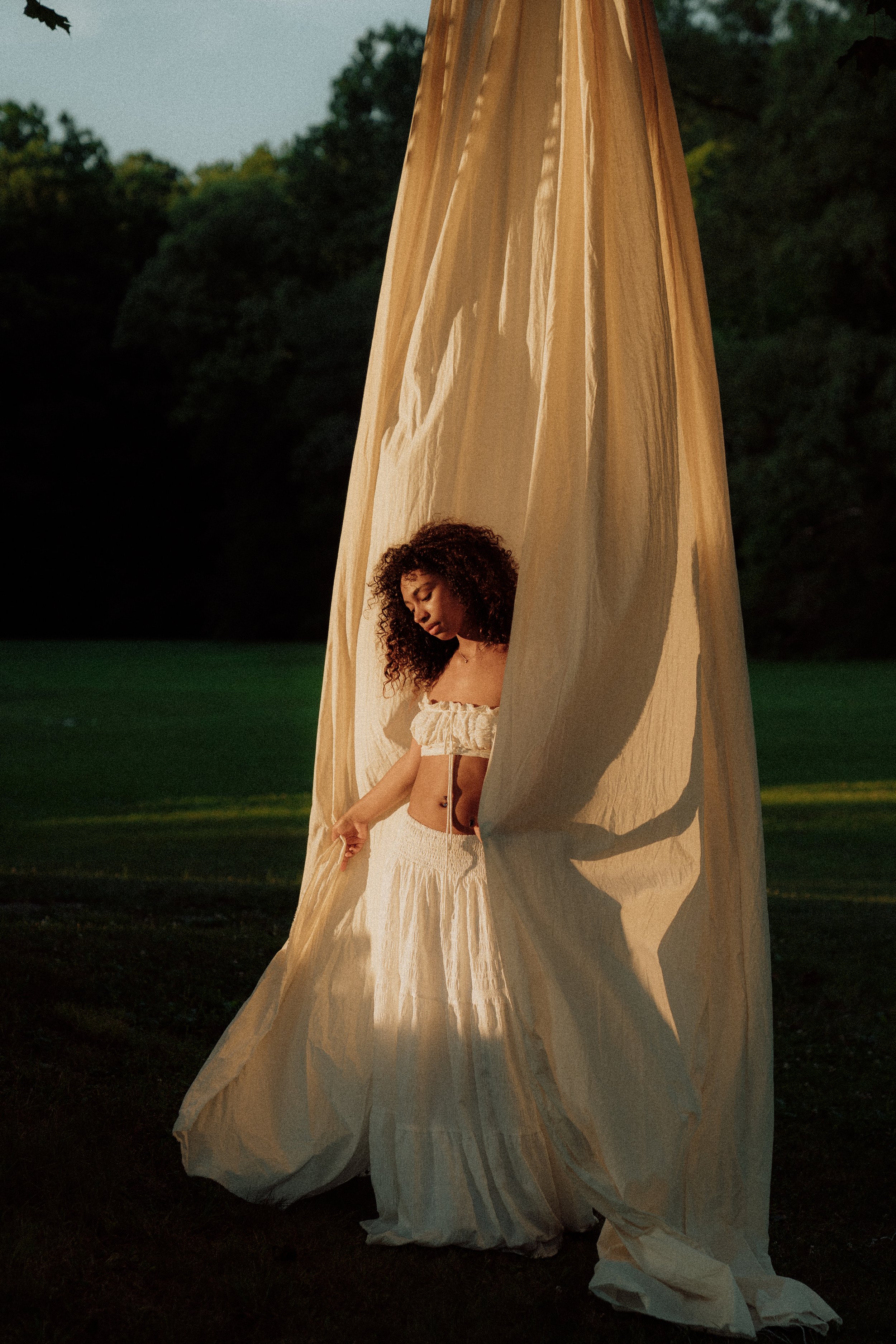 A woman with curly hair stands outdoors at dusk or dawn, partially concealed behind a large flowing beige fabric structure, with trees and open field in the background.