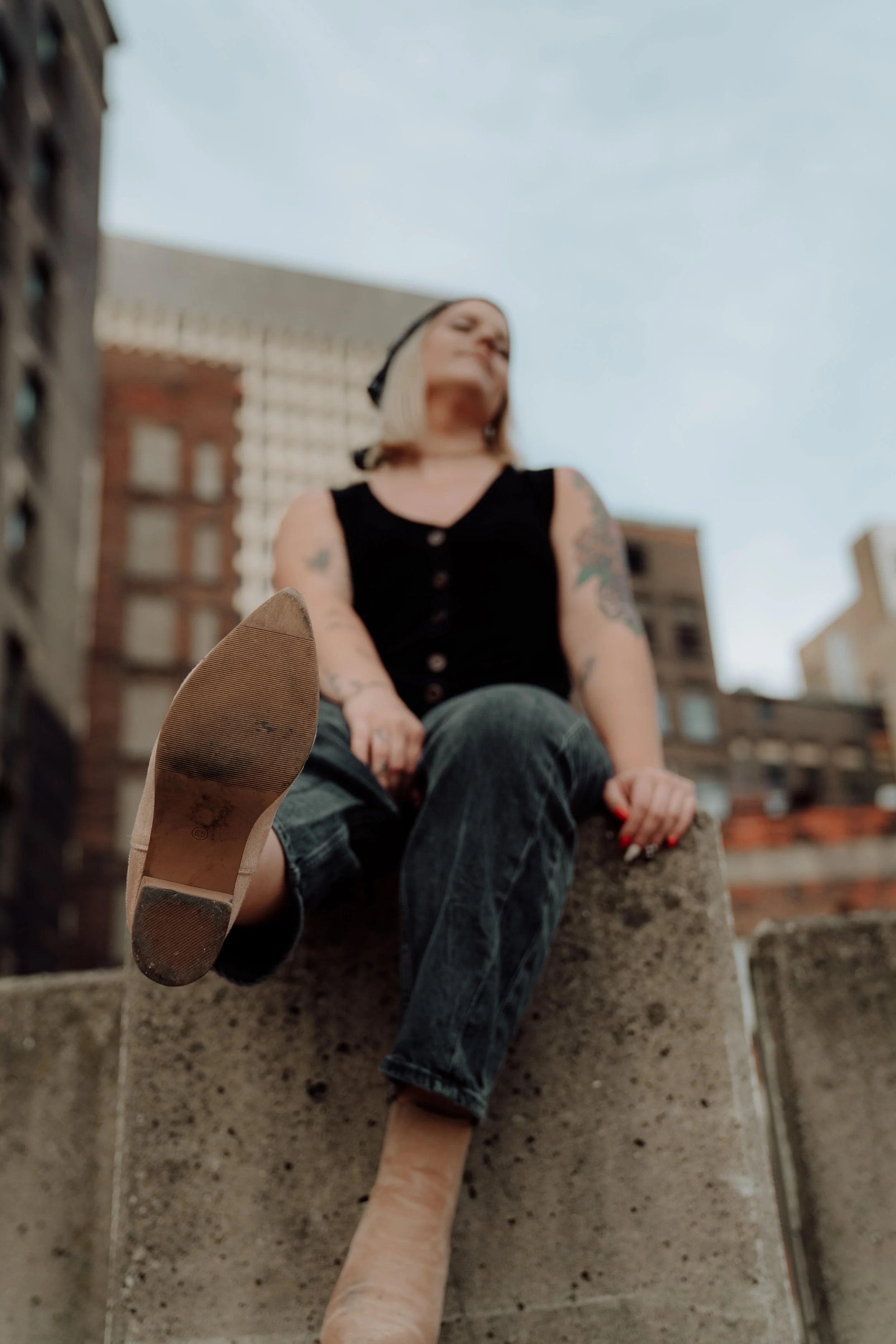 A woman with blonde hair, tattoos on her arms, wearing a black sleeveless top and glasses, is sitting on a concrete ledge in an urban setting, with buildings in the background. She is looking away, and the photo is taken from a low angle focusing on 